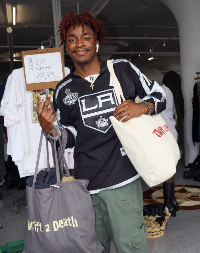 Person smiling and holding up a "V" sign while carrying two large tote bags in a clothing store. A sign behind them reads "$20 T-shirts".