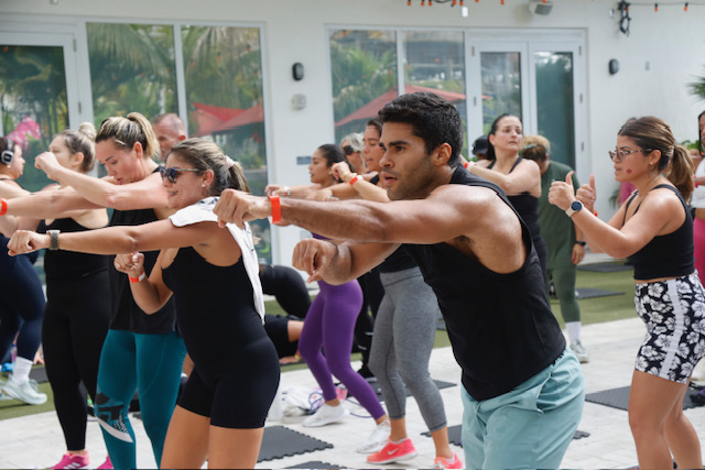 A group of people in workout attire participate in a synchronized fitness class, performing punch movements. They are outdoors, with exercise mats on the ground and a building in the background.