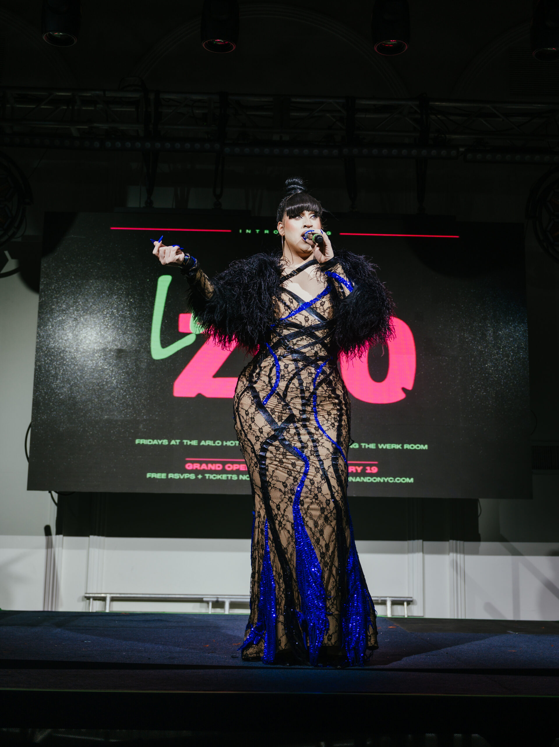 Person in a sheer black-and-blue dress with feathered sleeves stands on stage holding a microphone in front of a black screen with red and green text.