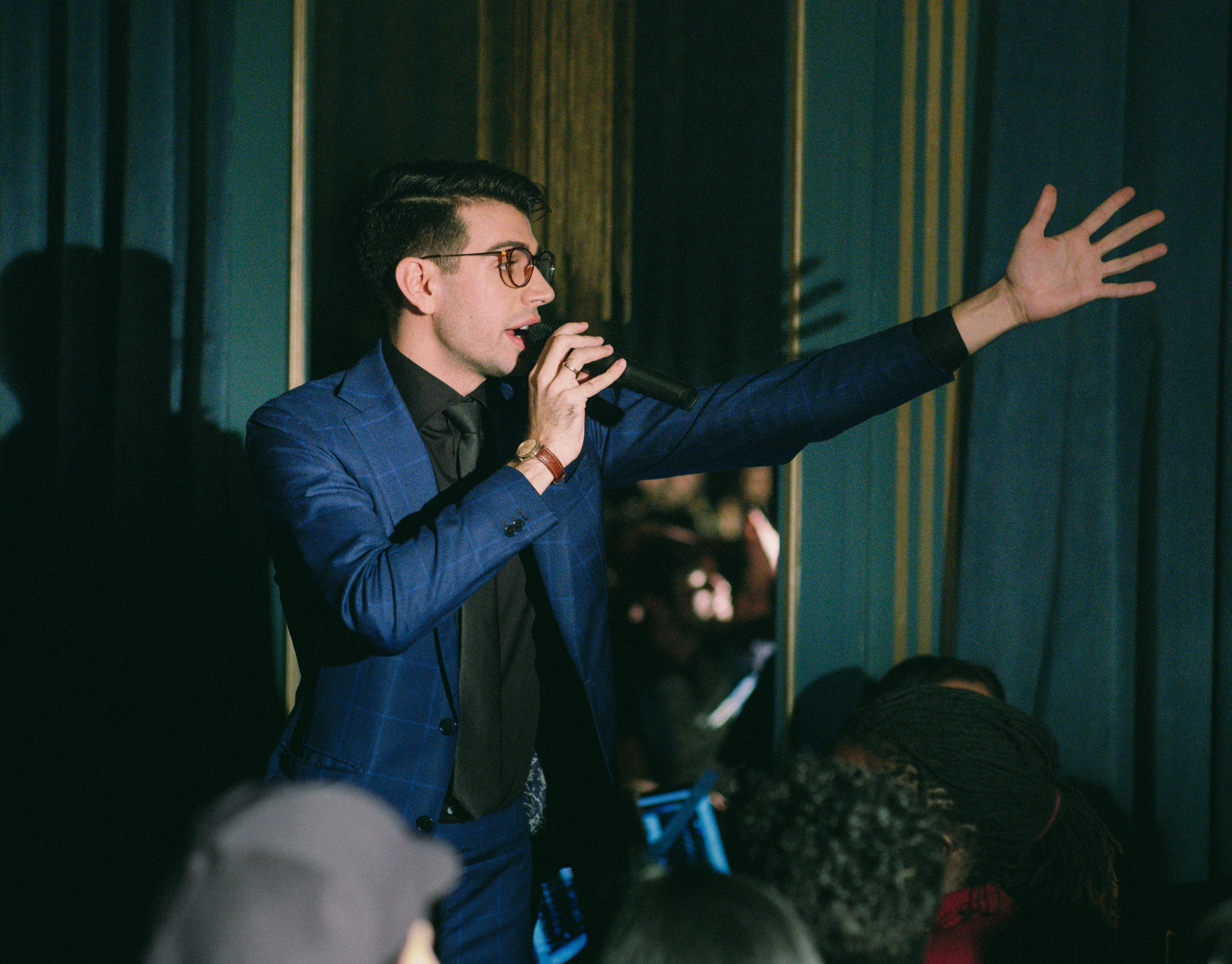 A person in a blue suit speaks into a microphone, gesturing with their right hand, in front of a dark curtain.