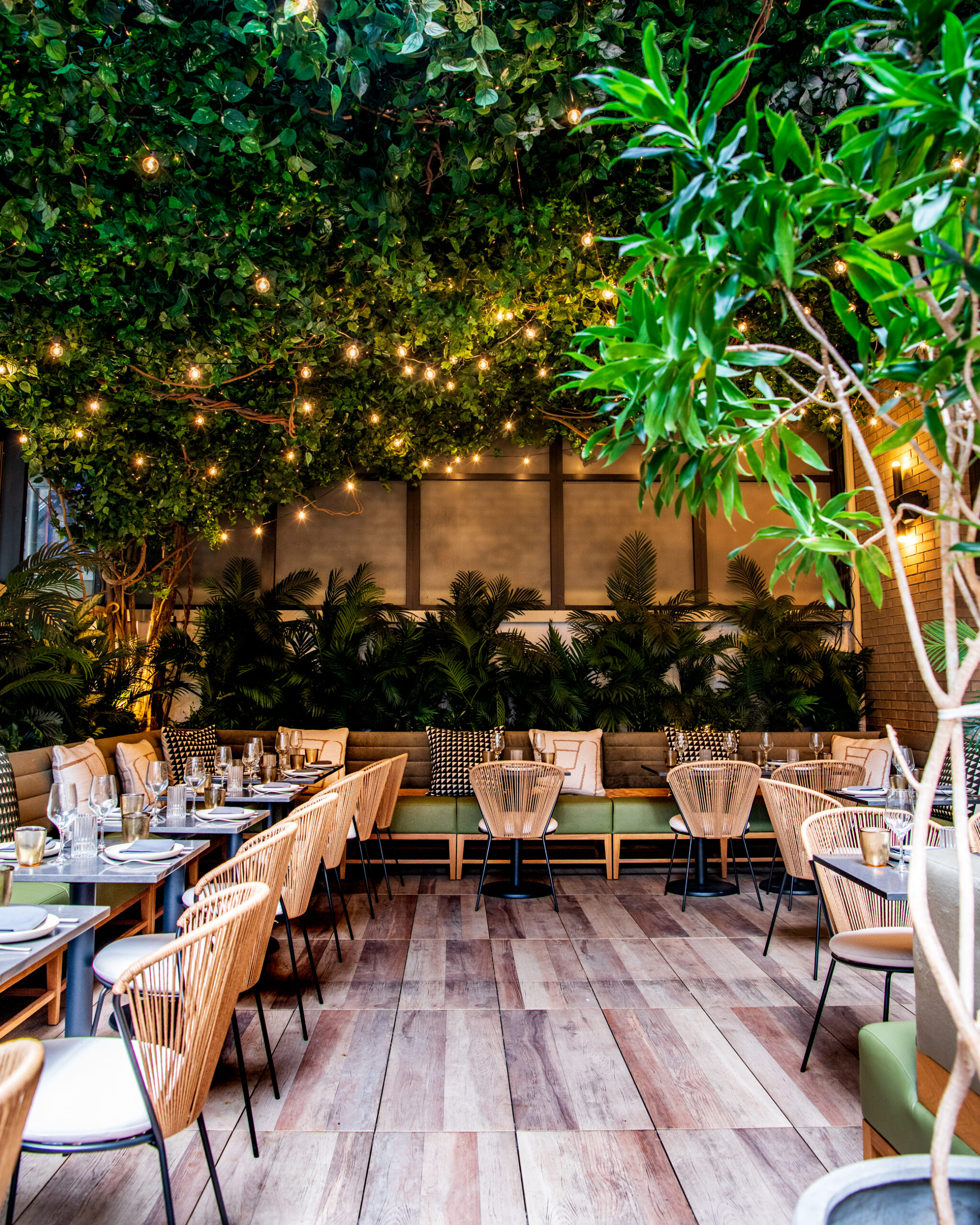 A cozy outdoor dining area with wooden tables and chairs, lush green plants, string lights overhead, and a wooden floor.