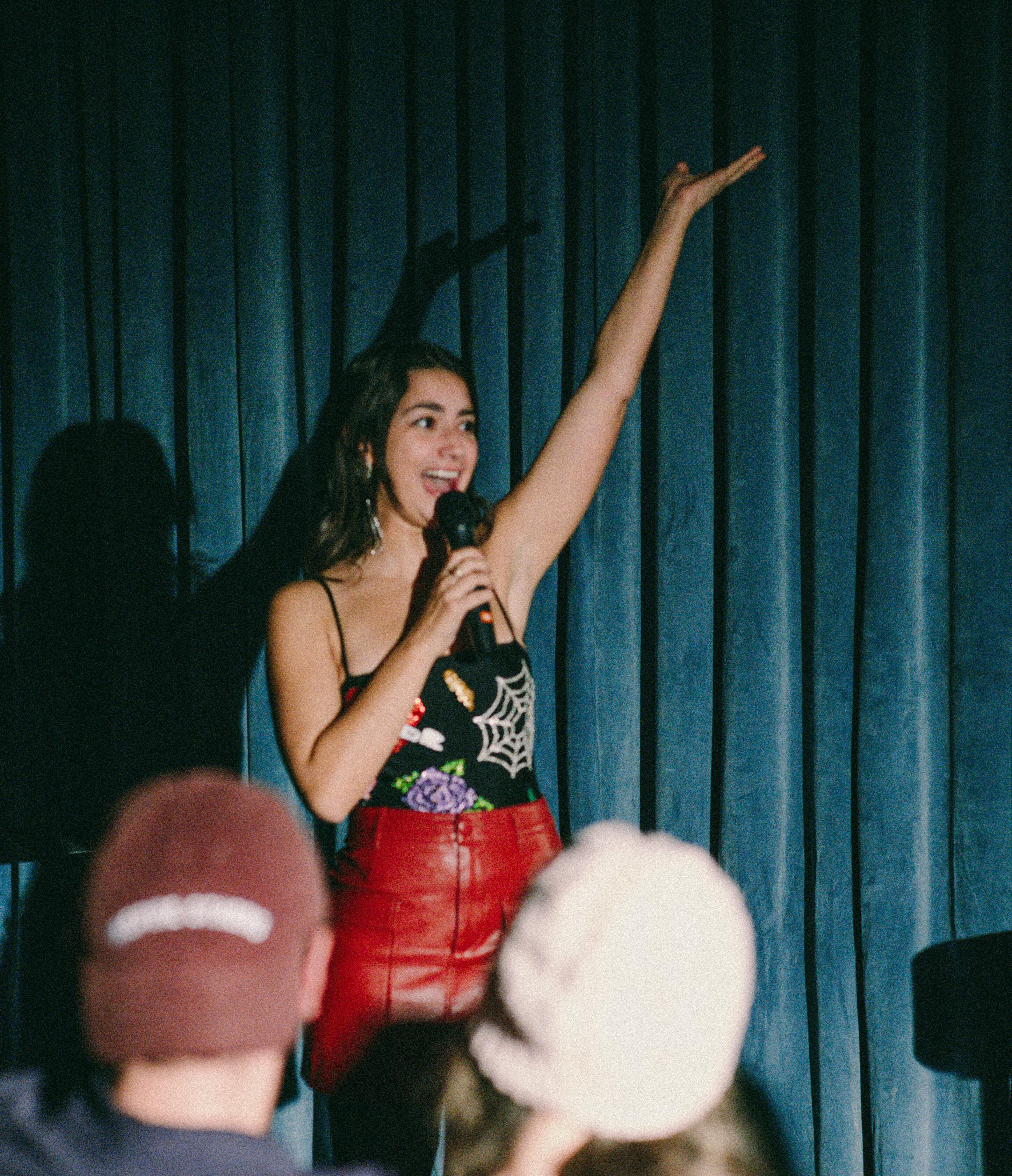 A woman stands on stage holding a microphone, raising one arm, and speaking to an audience in front of blue curtains.
