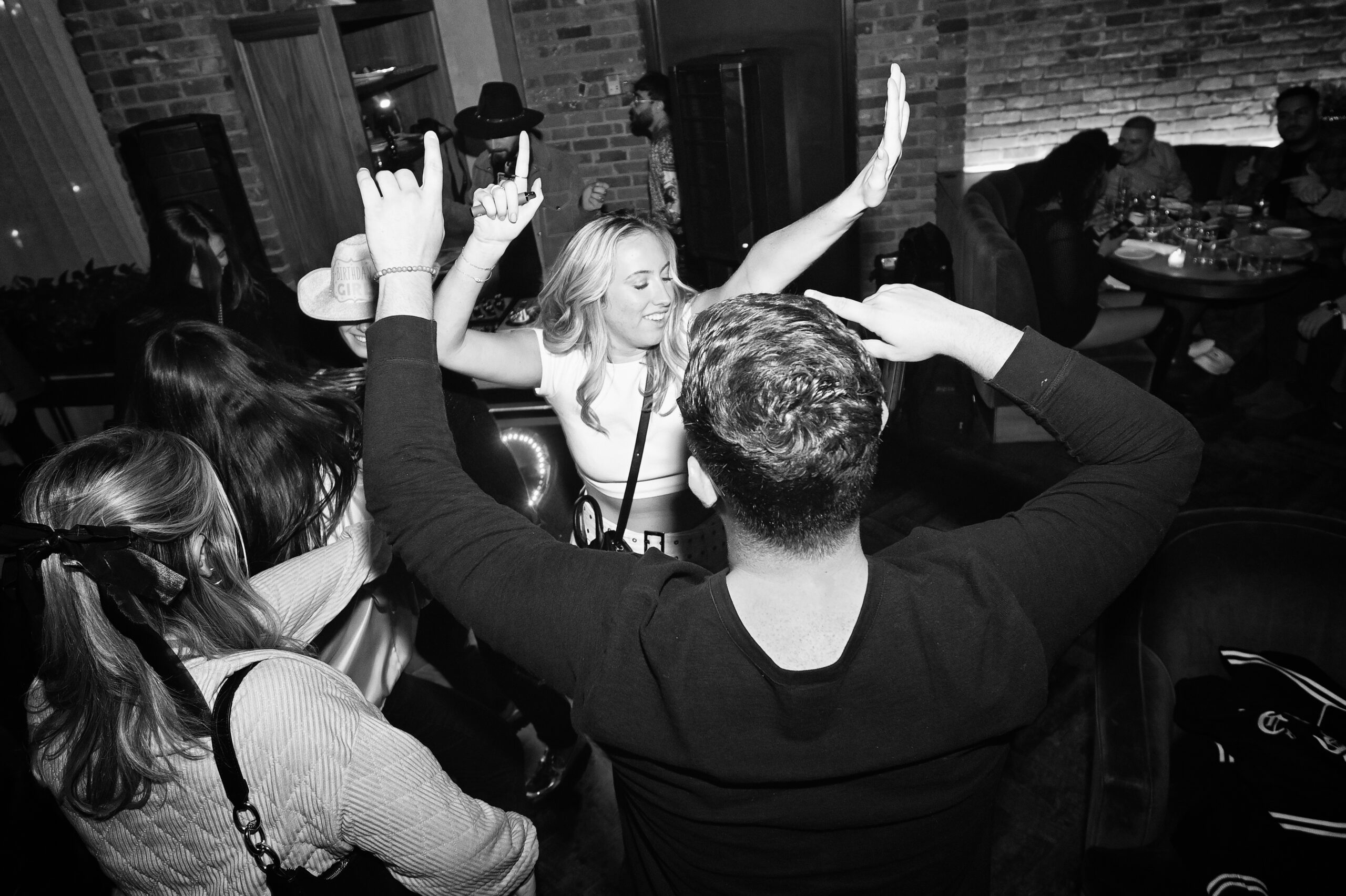 A group of people dancing energetically at a party in a dimly lit room with brick walls.