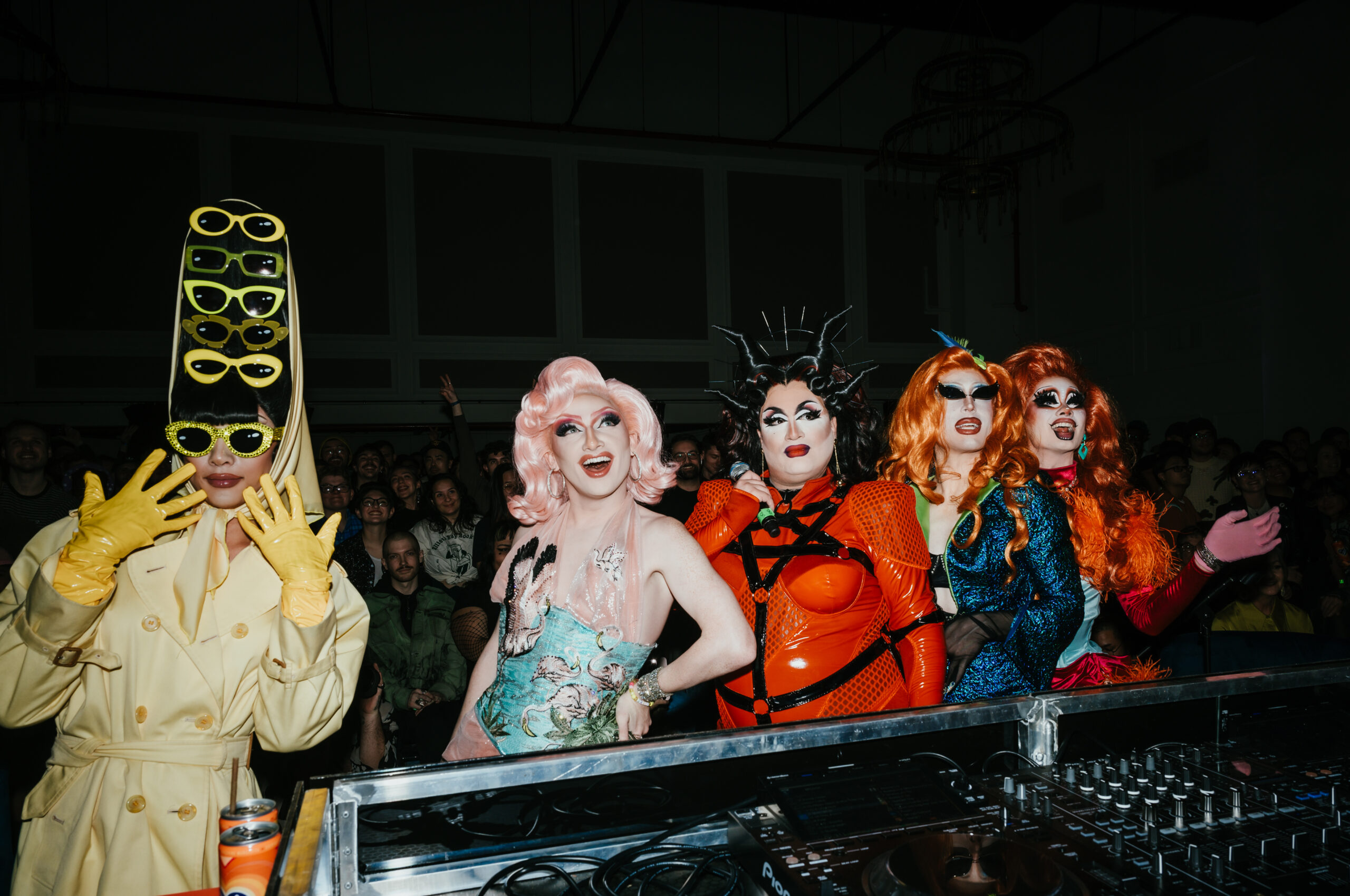 Five drag performers in colorful, elaborate costumes and wigs stand behind a DJ set in a dark venue with an audience in the background.