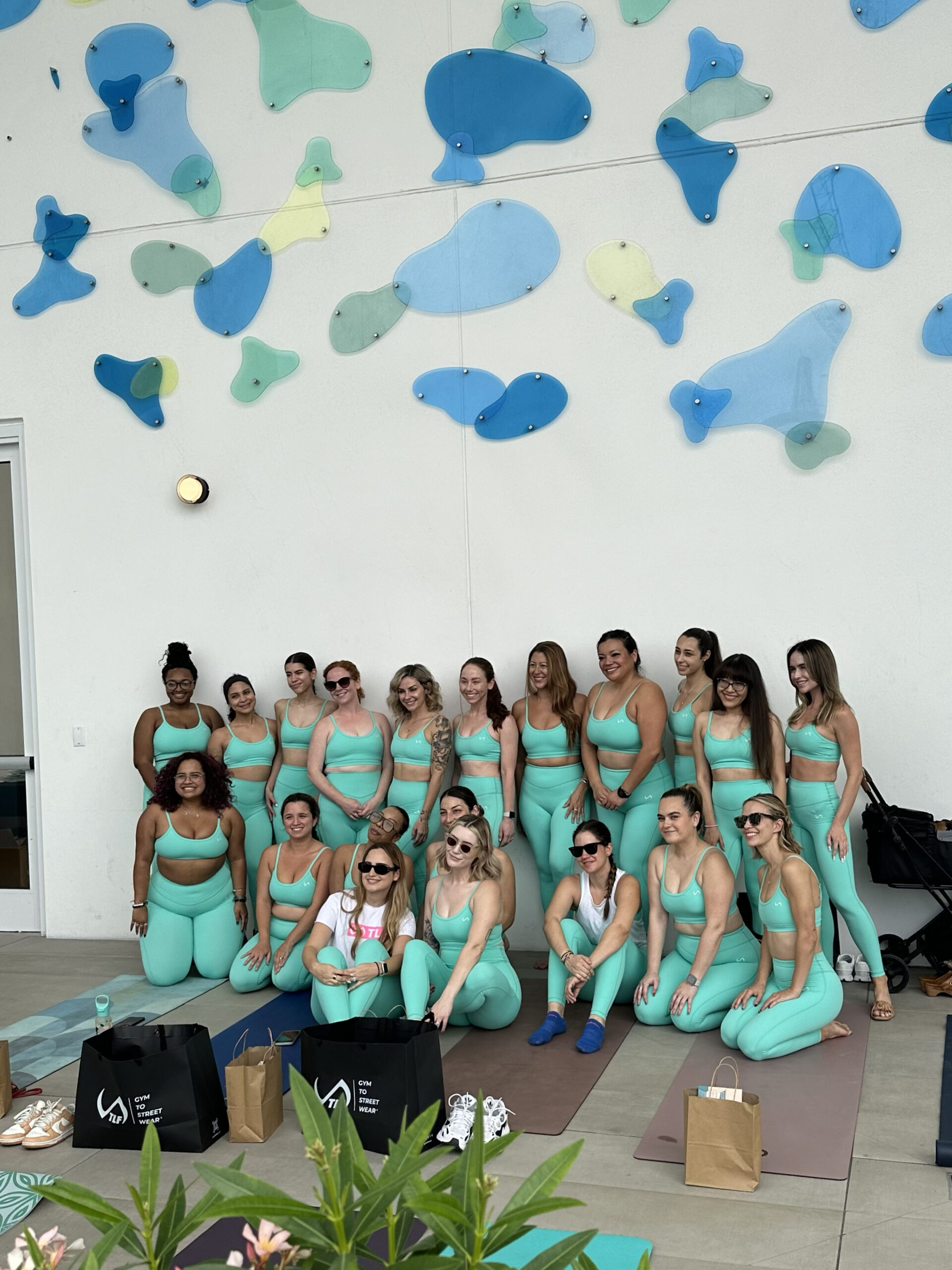 A group of people in matching turquoise workout attire posing together on yoga mats indoors. Wall in the background is decorated with abstract, colorful shapes. Shopping bags are in the foreground.