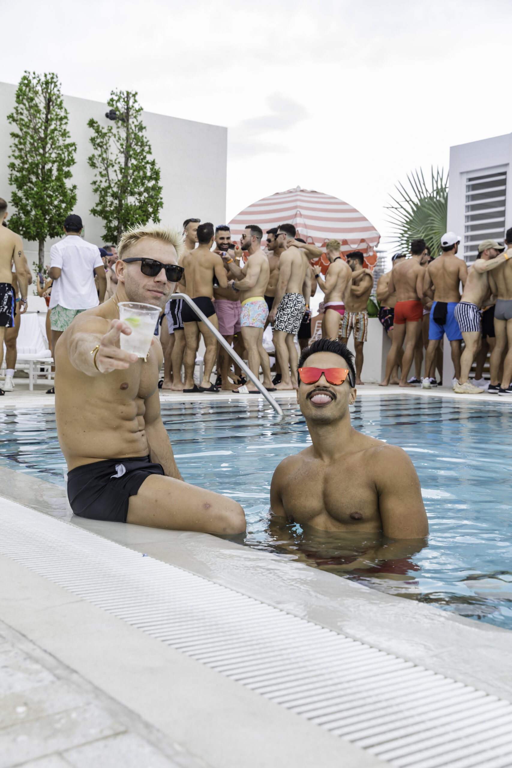 Two men are in a pool at a rooftop party. One man, wearing sunglasses, points towards the camera while holding a drink. The background shows other people socializing near the pool.