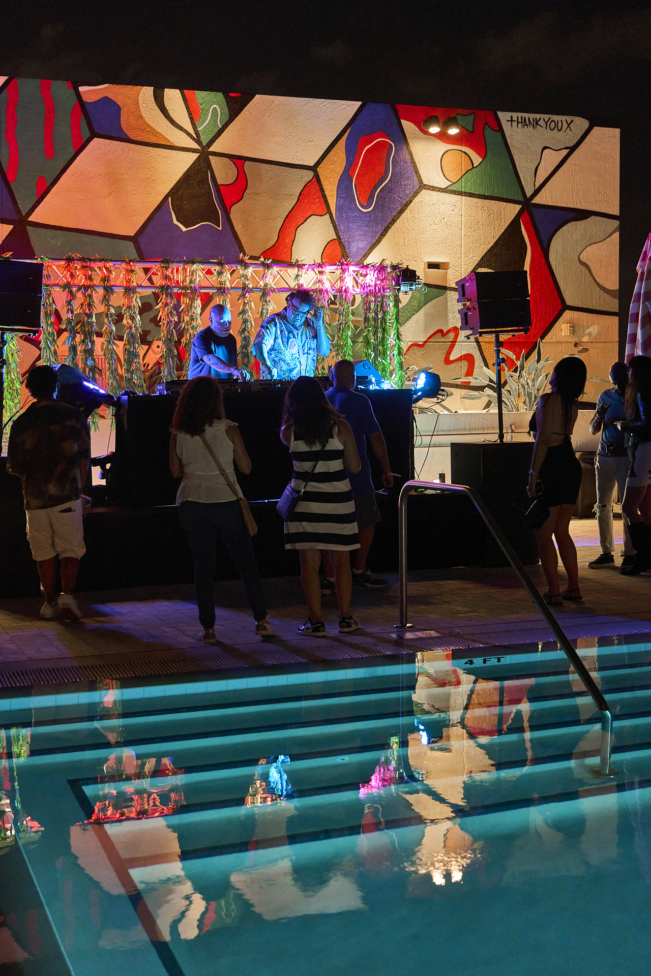 People gather near a pool under colorful lighting, facing a DJ booth with two DJs playing music. A vibrant mural is on the wall behind the DJs.