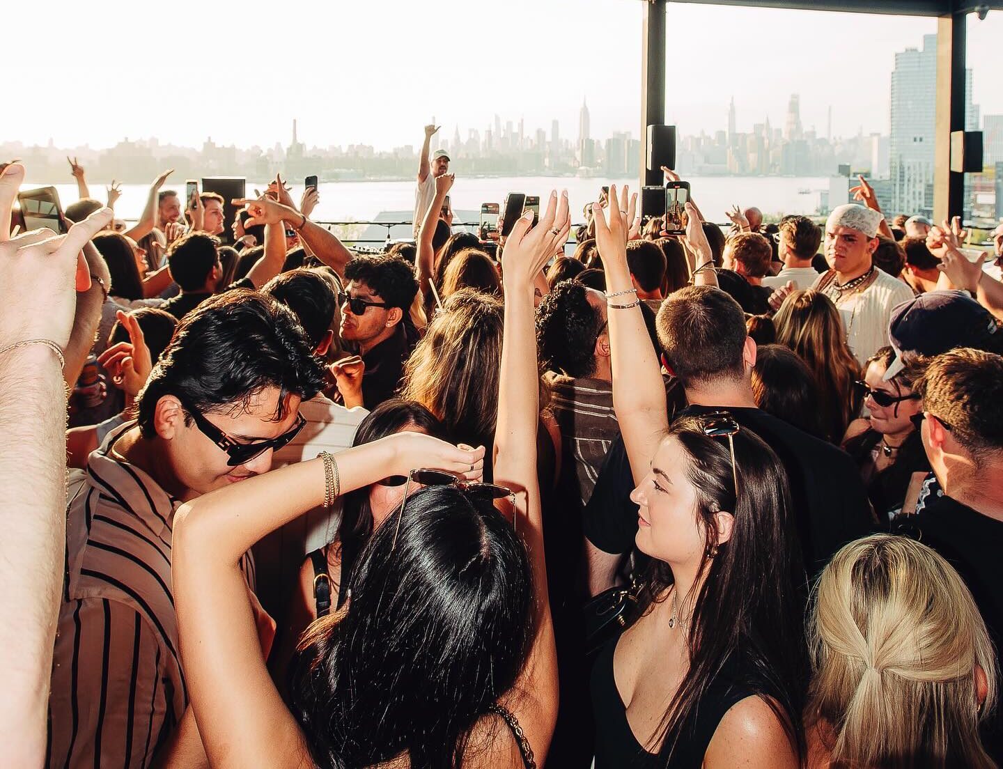 A crowded group of people are dancing and raising their hands at an outdoor rooftop party, with a city skyline visible in the background.