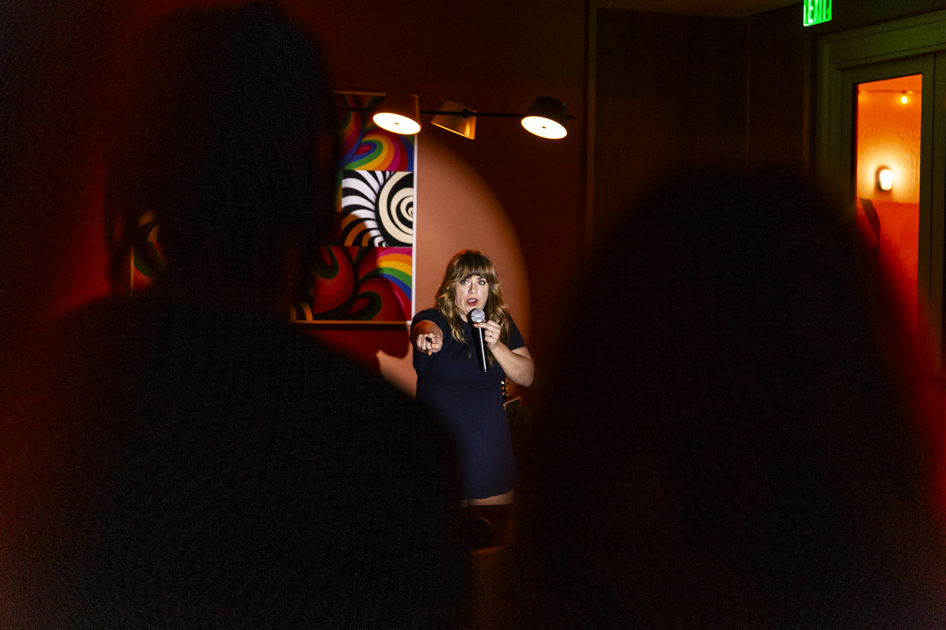 A woman performs stand-up comedy on stage, holding a microphone and pointing towards the audience. The room is dimly lit with abstract artwork on the wall.