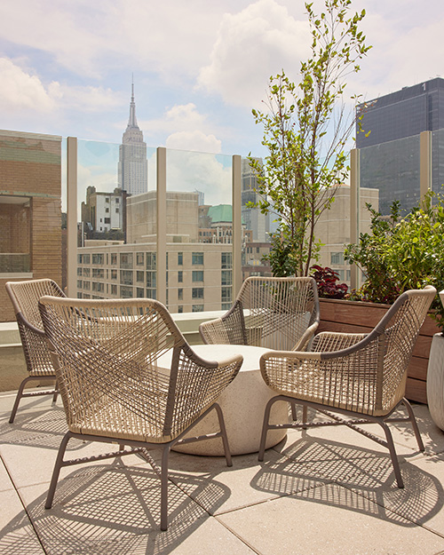 Rooftop seating area with four wicker chairs surrounding a round stone table, overlooking city buildings and the Empire State Building in the background.