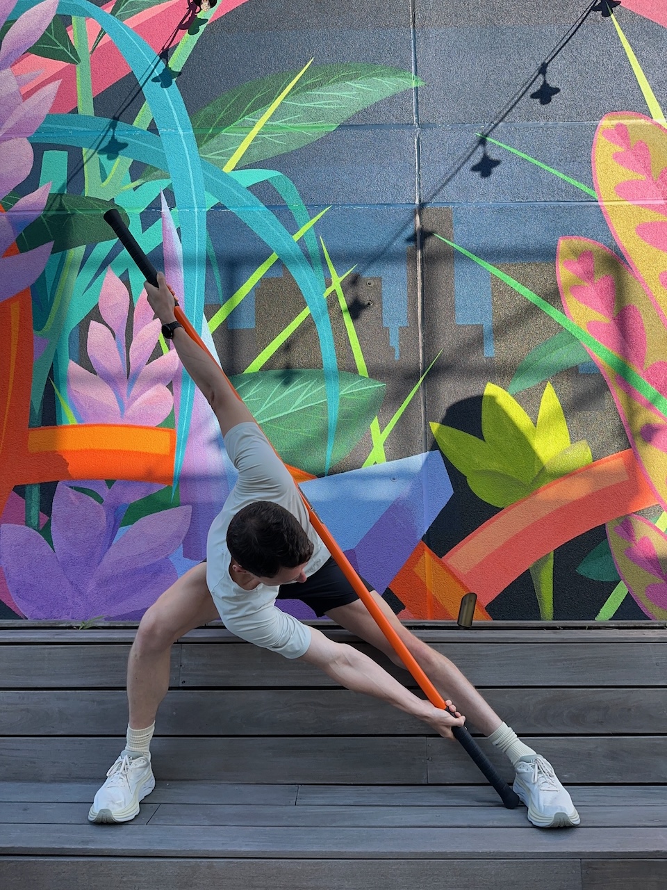 A person in athletic wear performs a stretch with an exercise bar against a colorful mural backdrop featuring abstract plant designs.