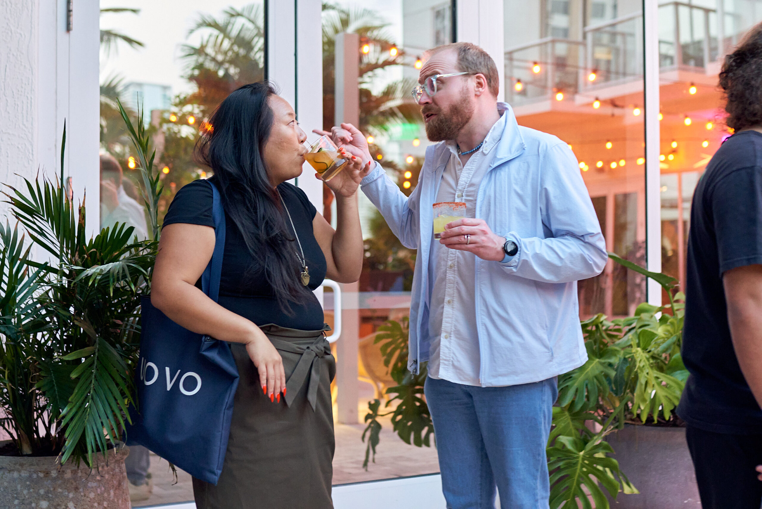 Two people stand outside a building with string lights, engaged in conversation while holding drinks. One person has a black tote bag on their shoulder. Plants can be seen in the foreground.