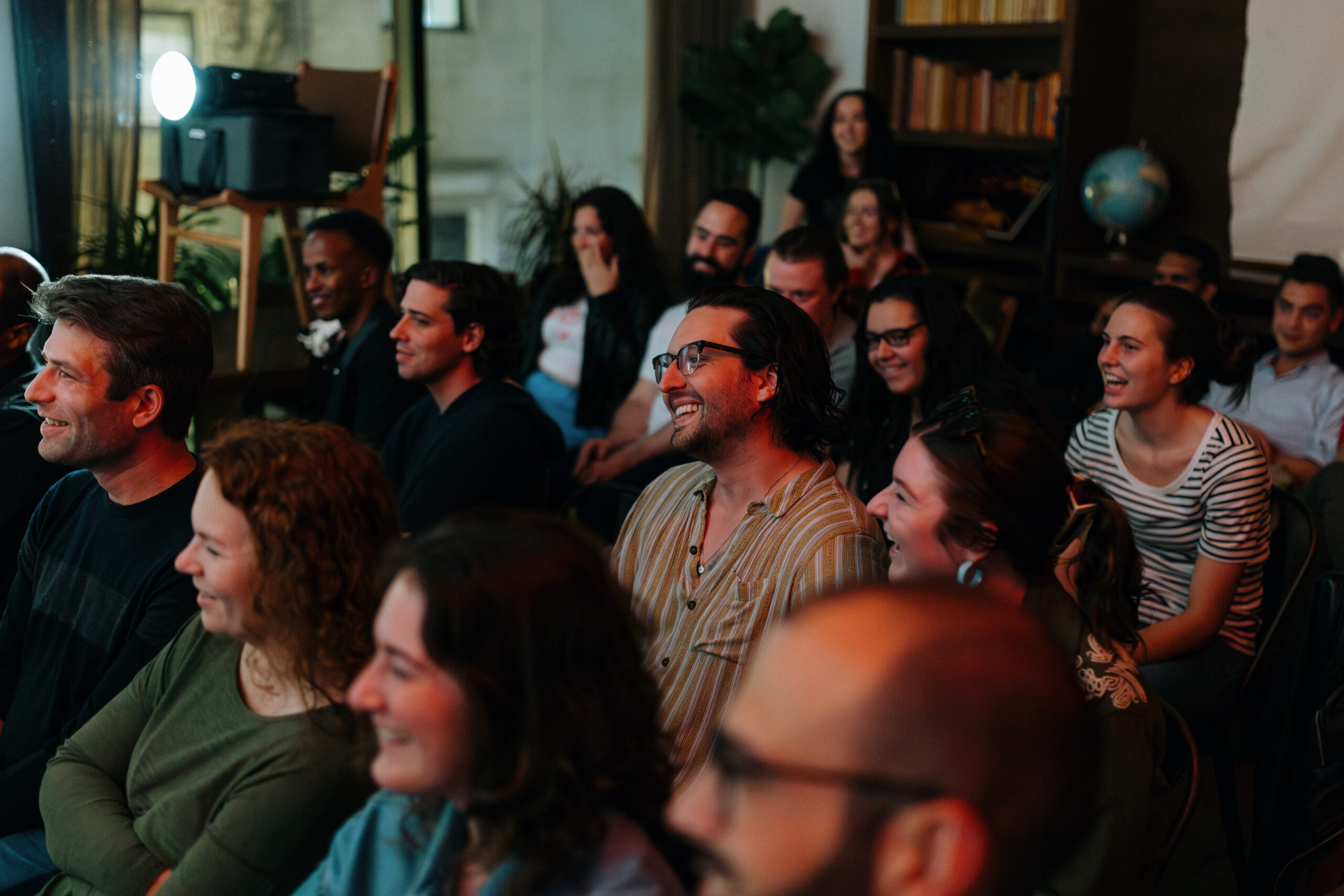 A group of people sitting closely together in a room, smiling and laughing while facing forward. A projector and bookshelf are visible in the background.