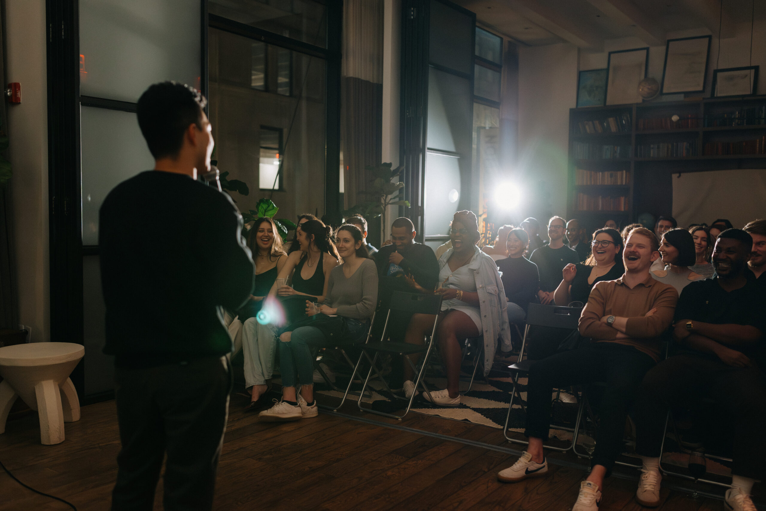 A person is speaking into a microphone in front of a seated audience in a well-lit room with large windows and bookcases in the background.