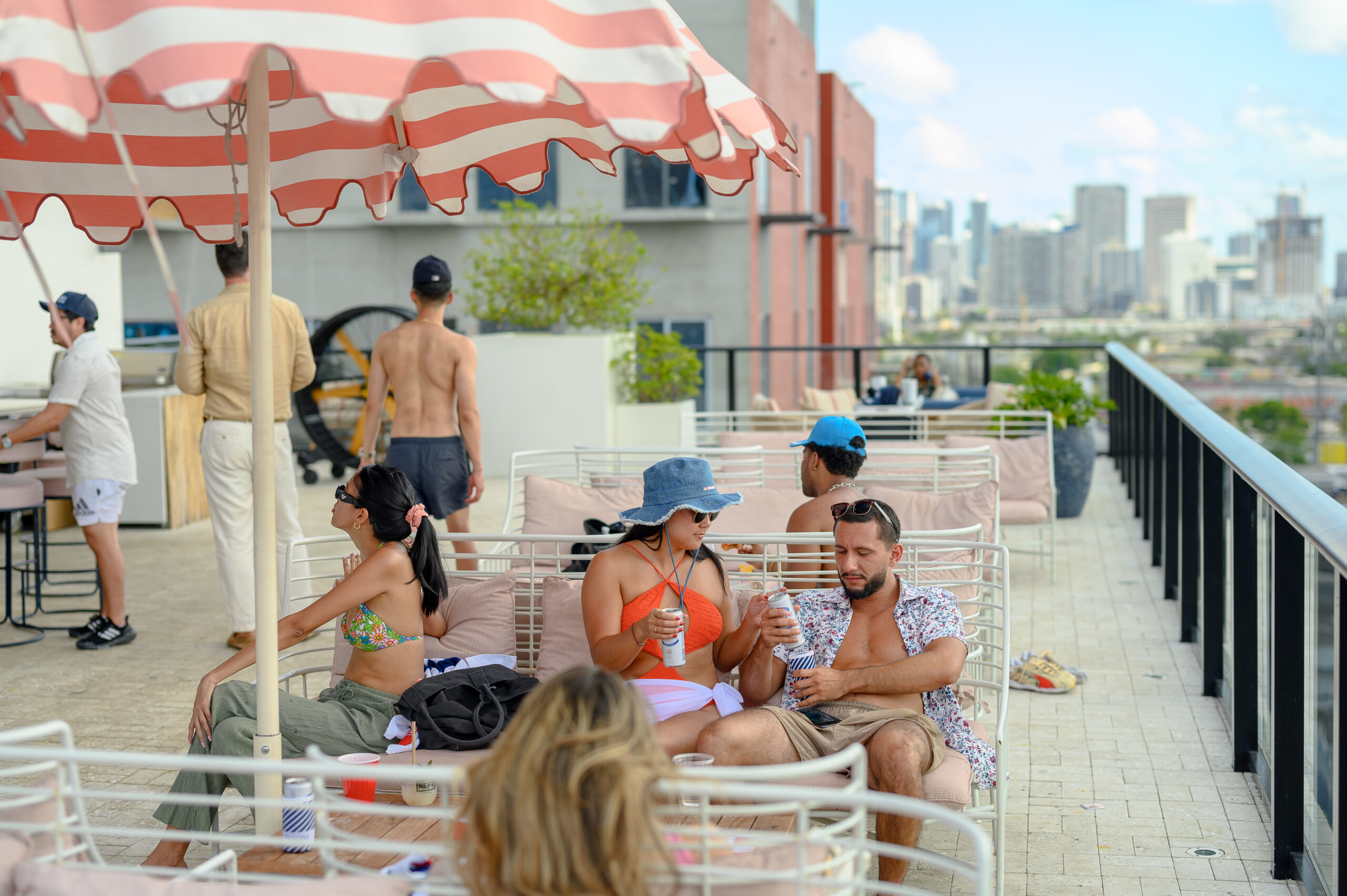 People relaxing and socializing on a rooftop terrace with city views. Some are under striped umbrellas, while others enjoy drinks and conversation.
