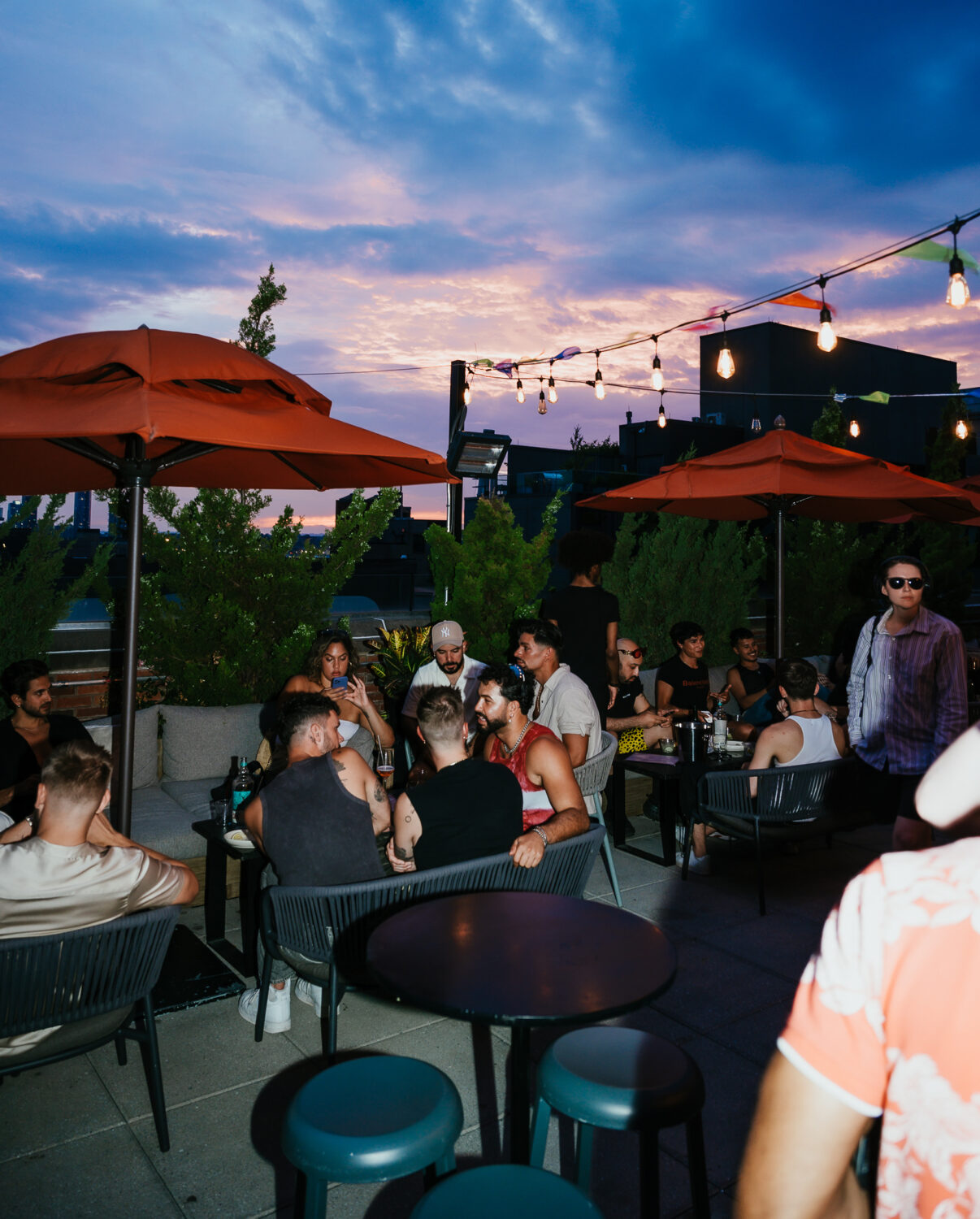 A group of people enjoy an evening at Arlo SoHogathering on a rooftop patio with string lights and orange umbrellas, against a backdrop of a colorful sunset sky.