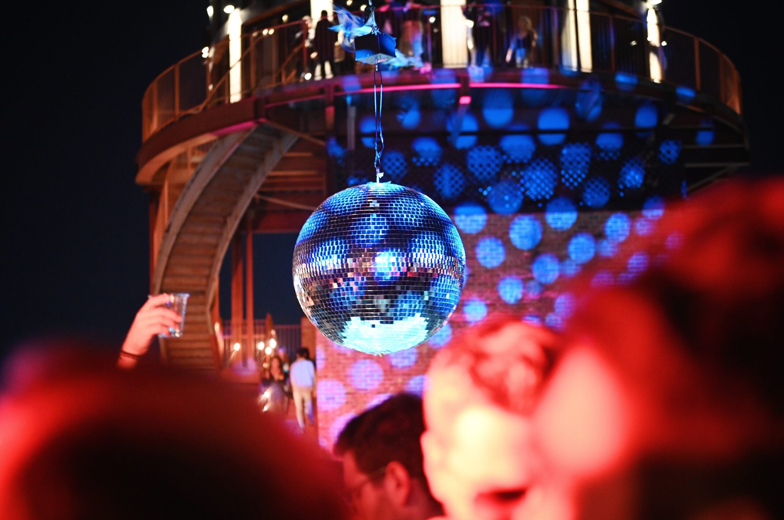 A reflective disco ball hangs in a crowded party scene with colorful lights. A person holds a drink to the left, and an elevated platform is in the background with people on it.