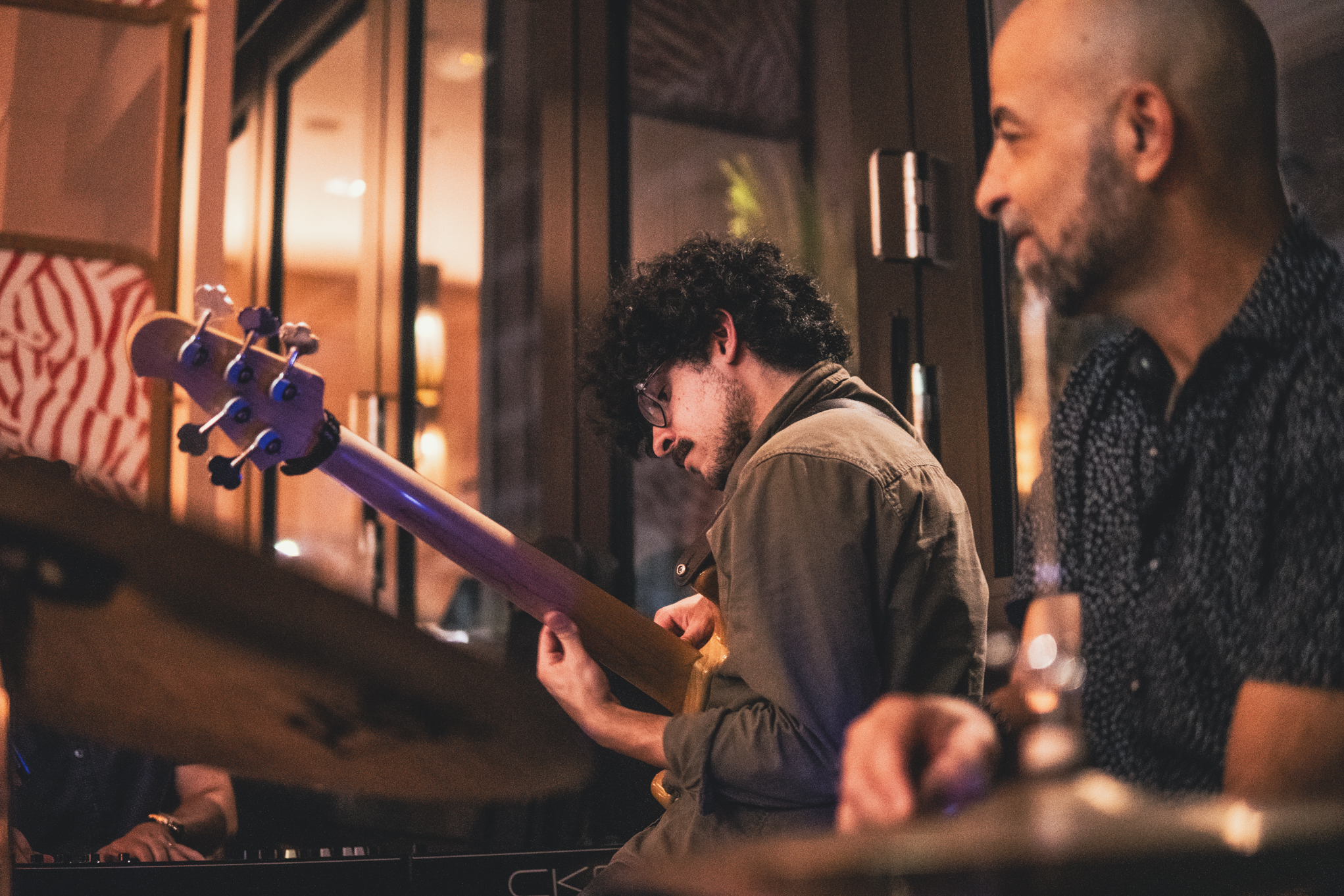 Two musicians perform; one plays a guitar and the other plays drums in a cozy indoor setting.