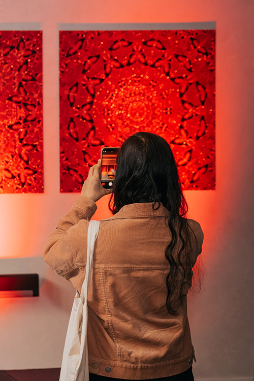 A person with long hair and a brown jacket takes a photo of two red patterned artworks on a wall using a smartphone.