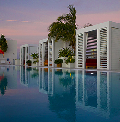 A serene outdoor pool with modern white cabanas and palm trees at sunset, reflecting pink and purple hues in the water.