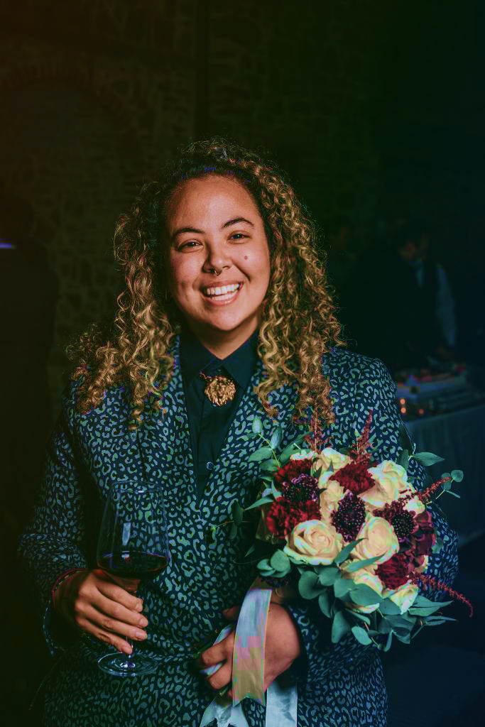 A person with curly hair smiles while holding a bouquet of flowers and a glass of red wine, wearing a patterned jacket, in a dimly lit room.