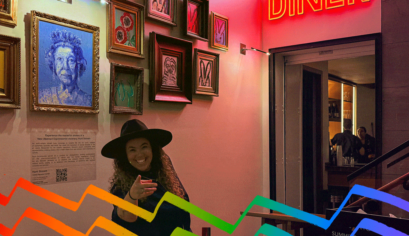A person wearing a hat points towards the camera while standing in front of an art wall next to a neon sign that reads "DINER." The reflection of another person is seen in the glass door nearby.