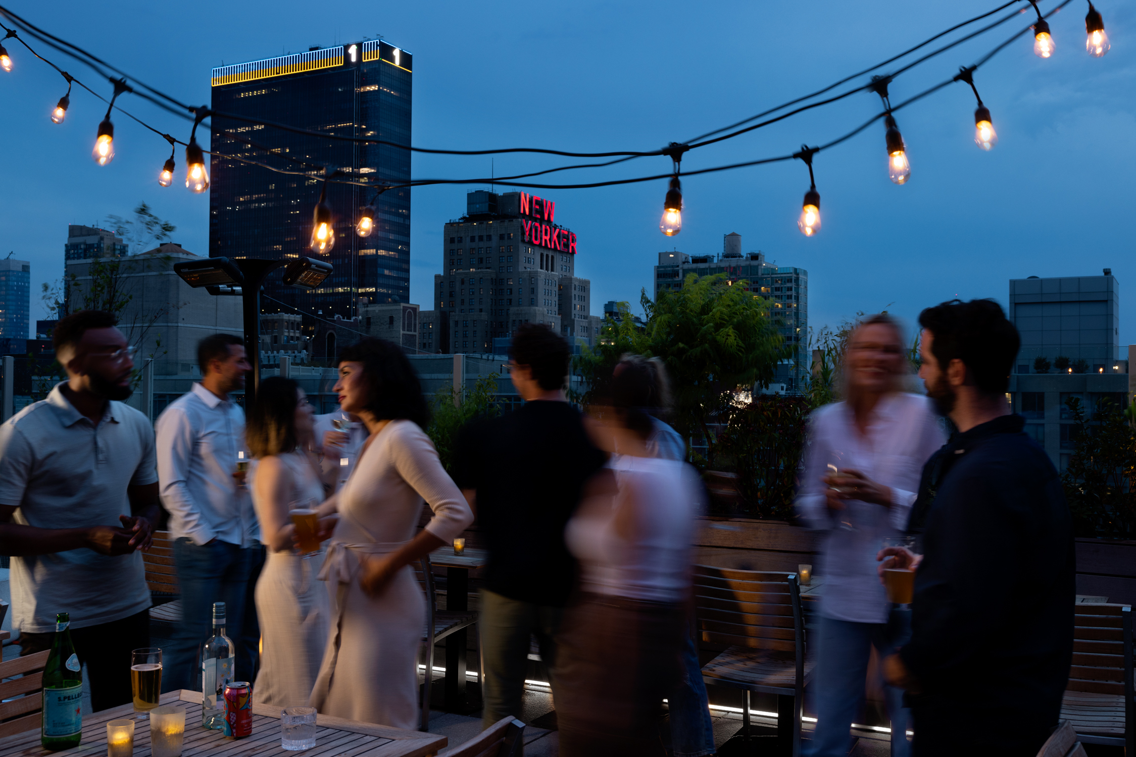 A group of people socializing on a rooftop at dusk with string lights overhead and city buildings, including one with a "New Yorker" sign, in the background.