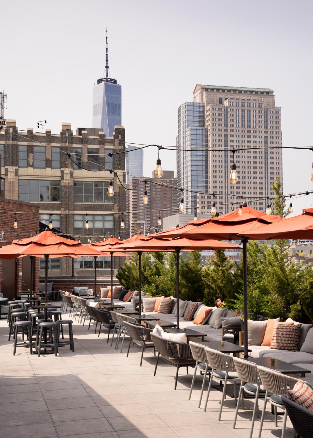 Rooftop patio with orange umbrellas, outdoor seating, and view of urban buildings, including a tall skyscraper in the background, under a clear sky.