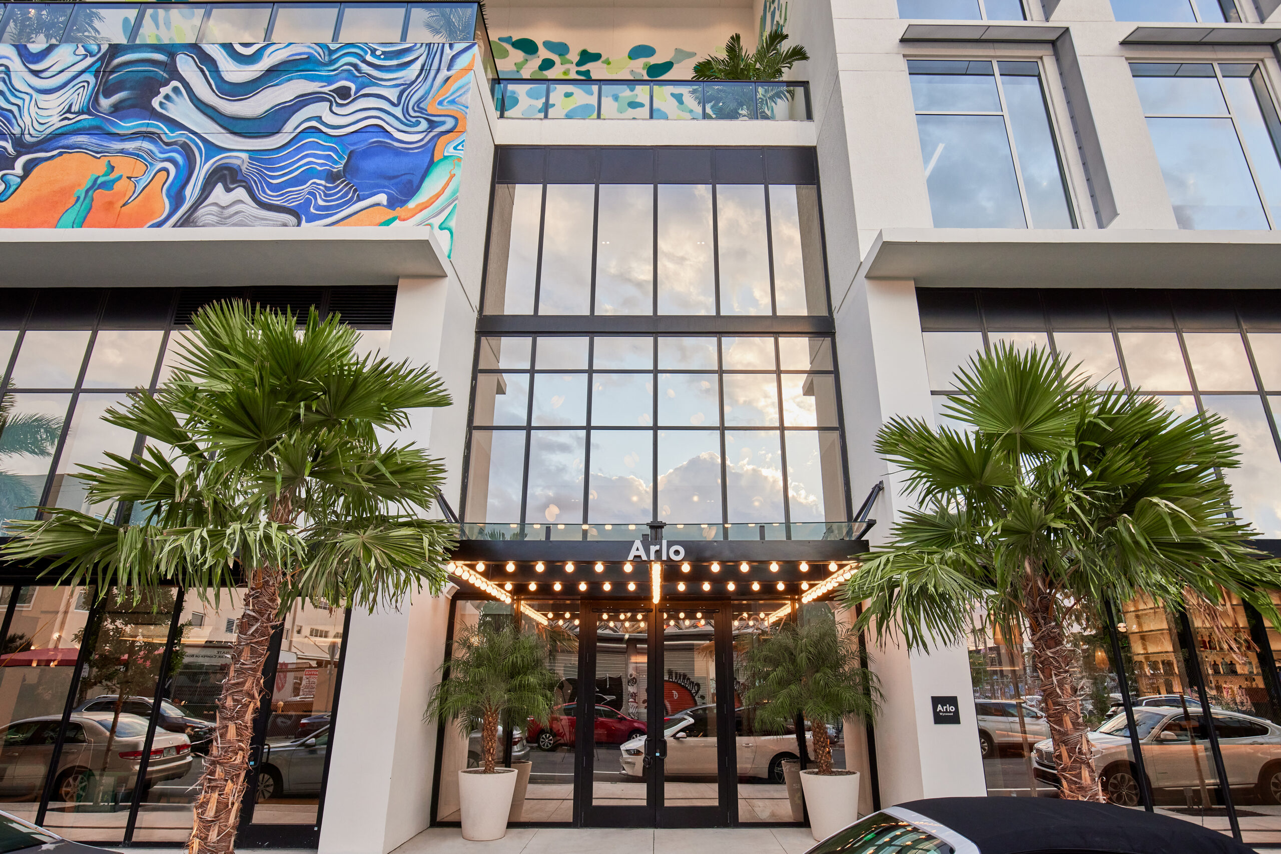 The entrance to the Arlo hotel with glass doors and a sign above. Palm trees in planters flank the doorway, and colorful murals adorn the building’s exterior.