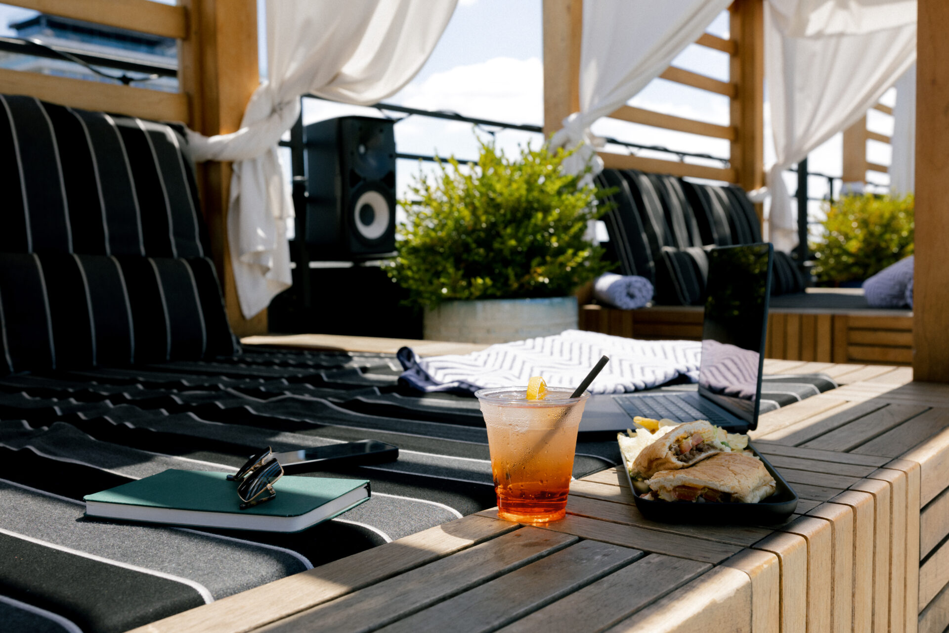 Outdoor lounge area with striped black and white cushions, a notebook, sunglasses, a drink with a straw and garnish, and a plate of food on a wooden surface, set against a sunny backdrop.