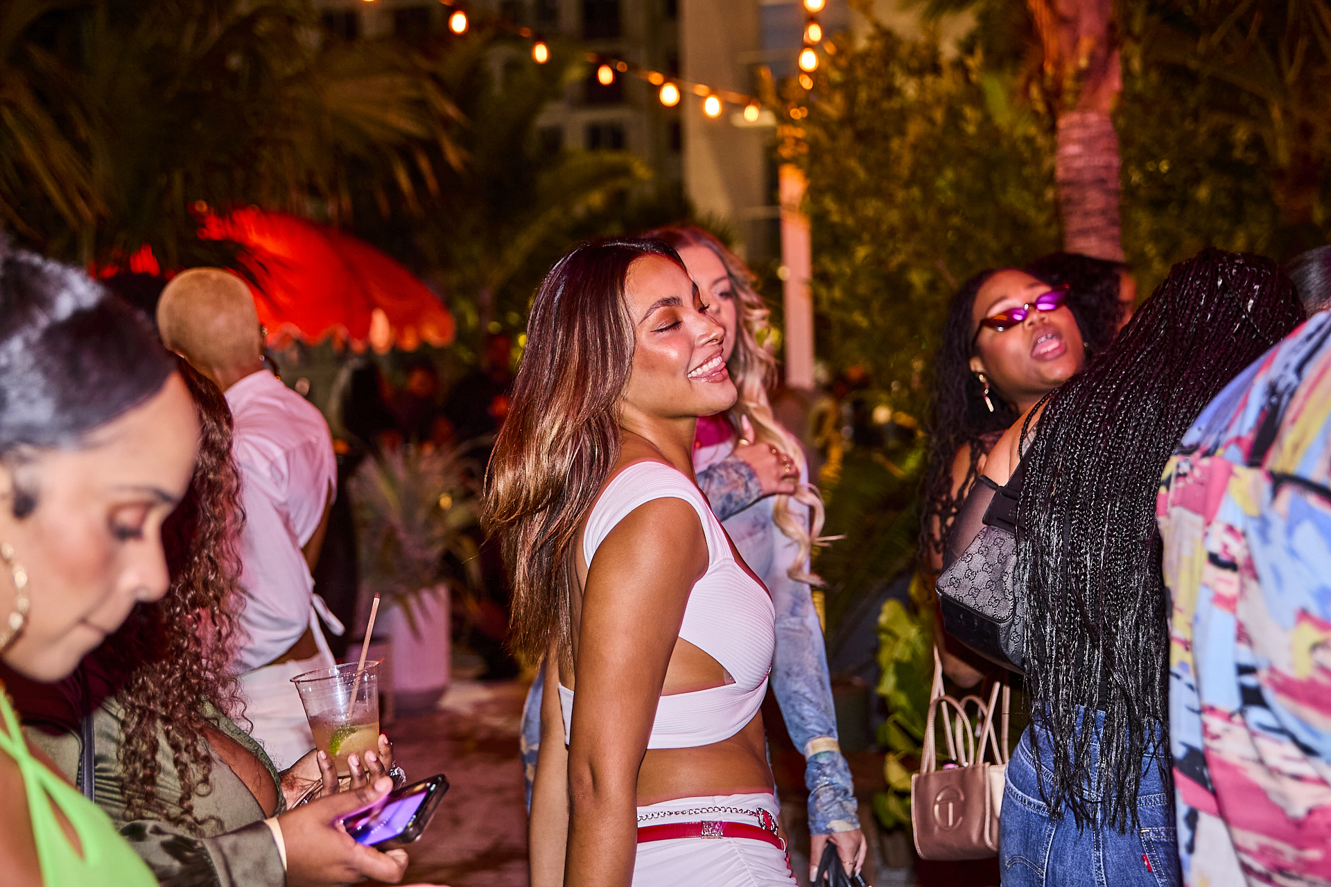 A group of people gathered at an outdoor nighttime event, some laughing and talking. One woman in the center is smiling, while others around her are engaged in various activities like checking phones.