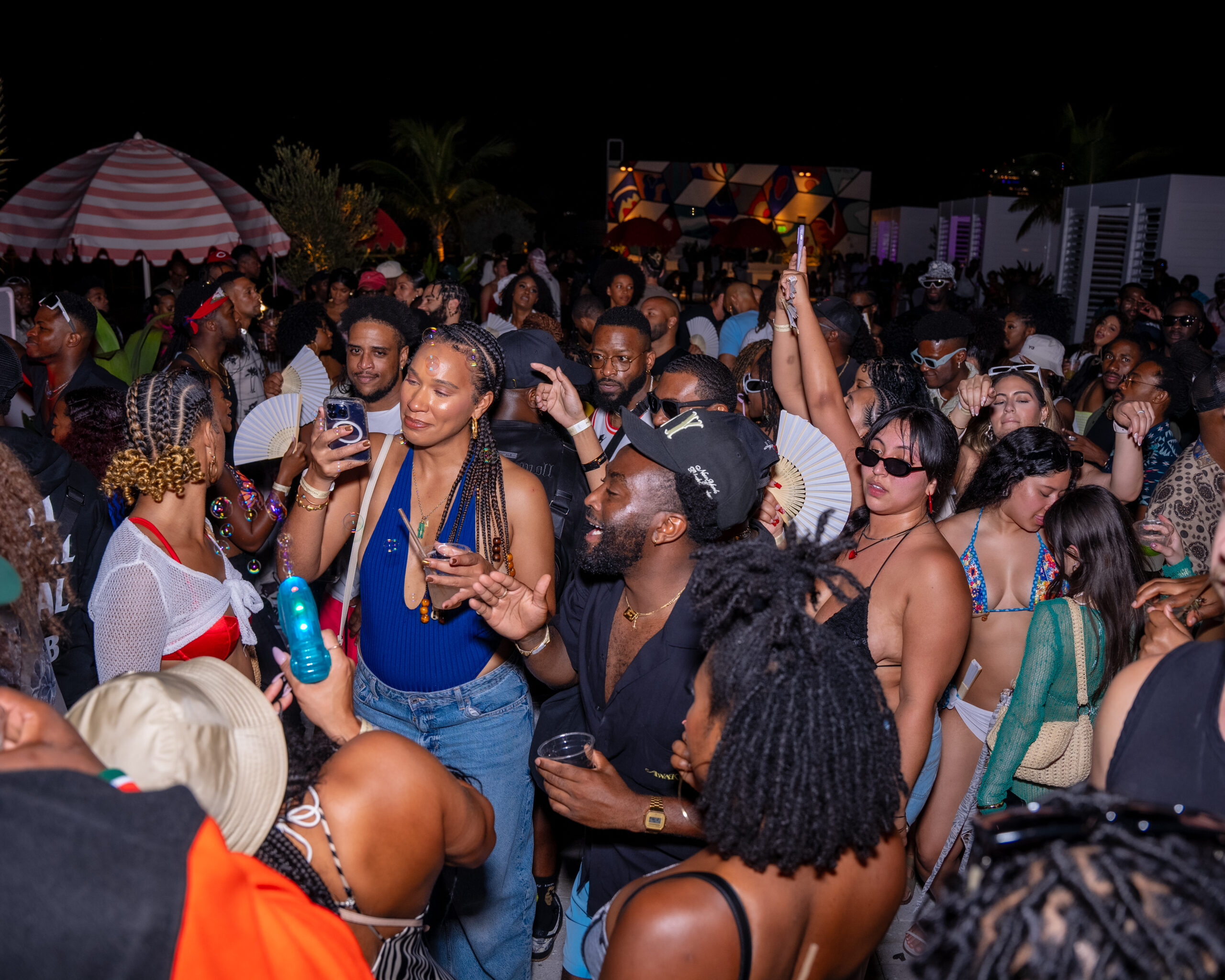 A large group of people enjoying a lively outdoor party at night, some holding drinks and phones, others dancing and chatting. The scene includes colorful decorations and a festive atmosphere.