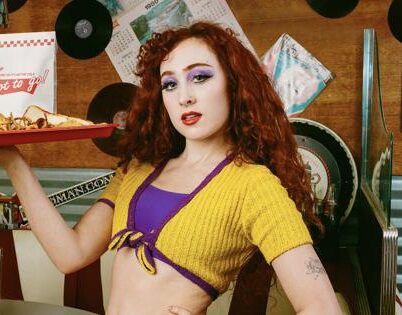 A woman with red curly hair and purple eye makeup holds a red tray with food in a retro diner setting. The tray contains a to-go bag, two baskets with food, and napkins. She is wearing a yellow cropped top.