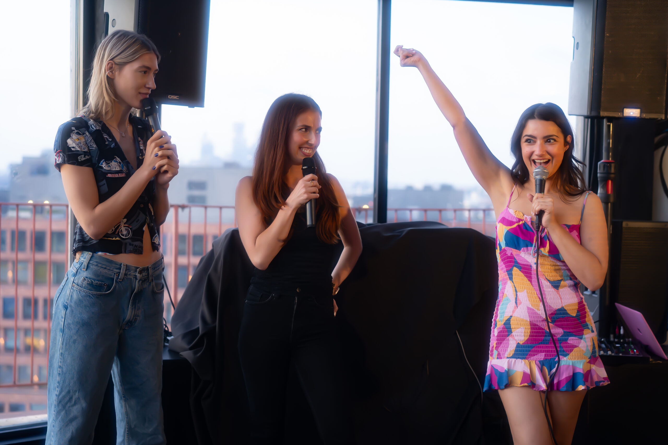 Three women are standing indoors with microphones. One woman is raising her arm enthusiastically while the other two look on and smile.