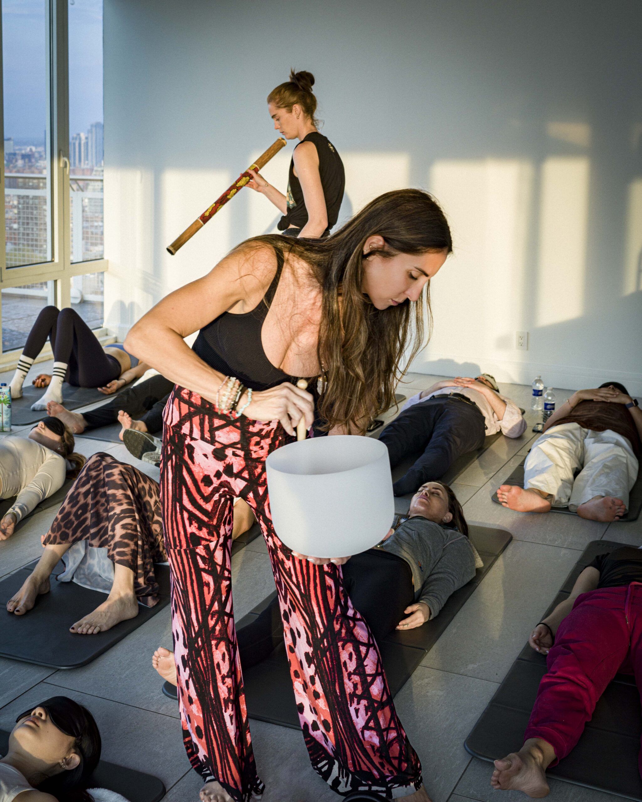 A woman plays a singing bowl while another plays a flute during a group relaxation session with participants lying on mats in a bright room.