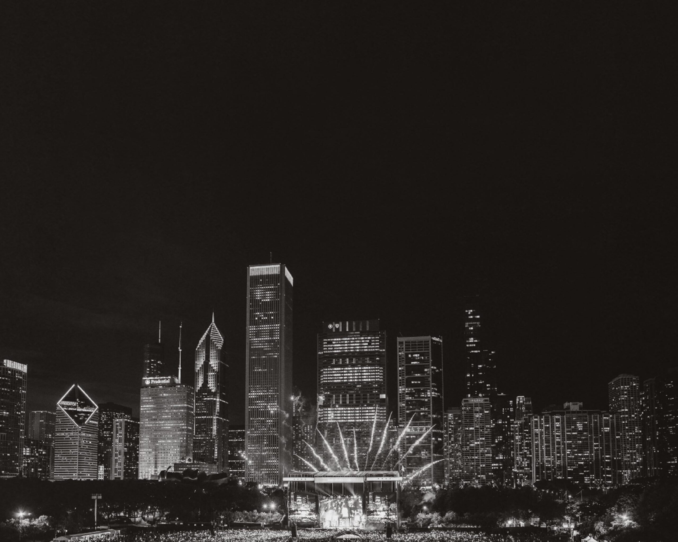 Black-and-white image of a cityscape at night featuring tall, illuminated buildings. Bright lights radiate from a central structure, creating a dramatic effect over a gathering of people below.