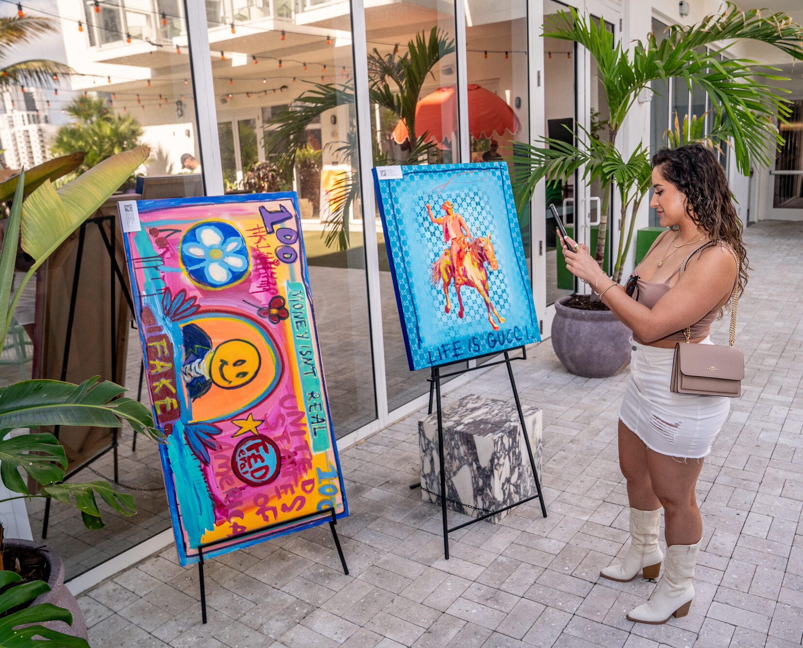 A woman in a white dress takes a photo of two colorful artworks displayed on easels at an outdoor gallery.