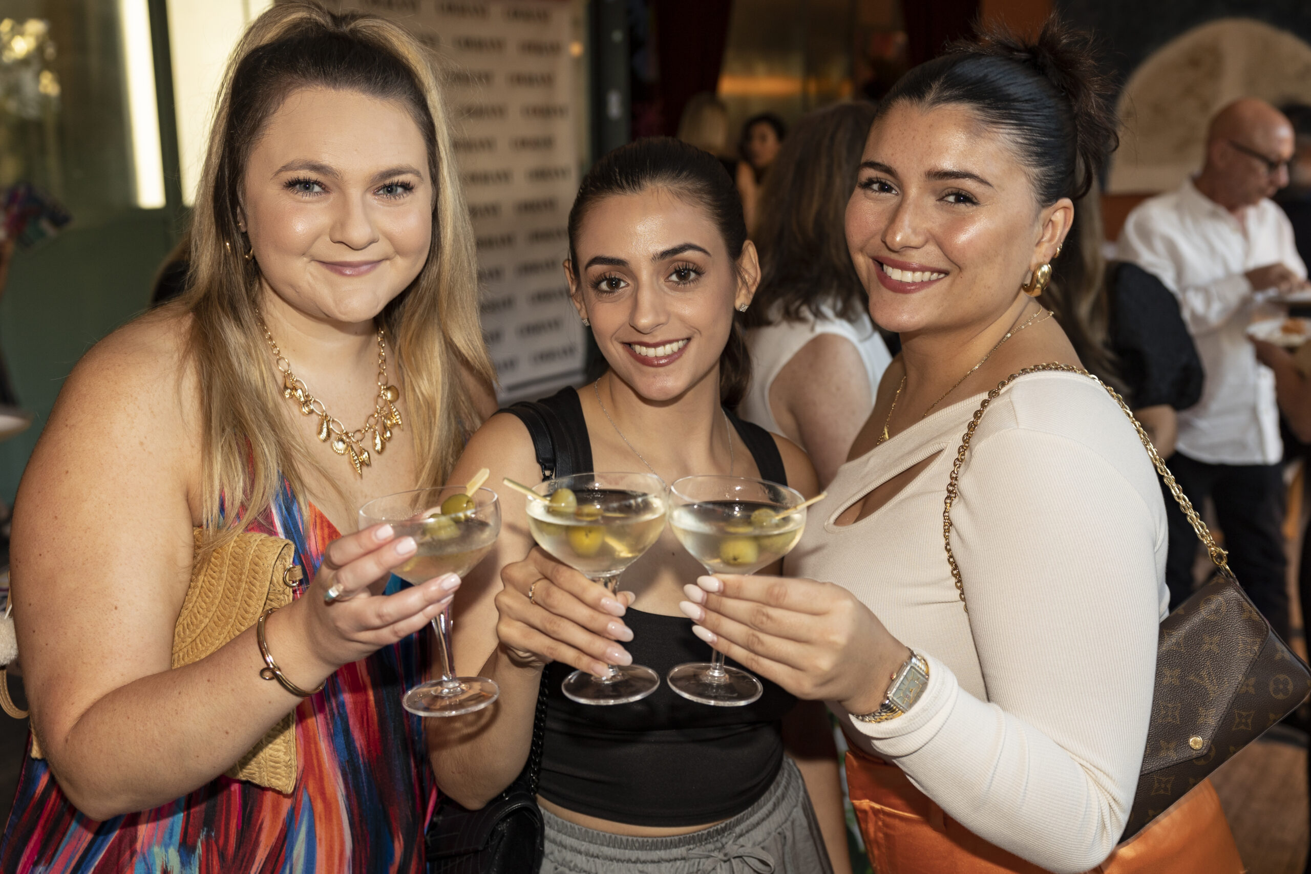 Three women holding martini glasses and smiling at the camera, while others mingle in the background at a social gathering.