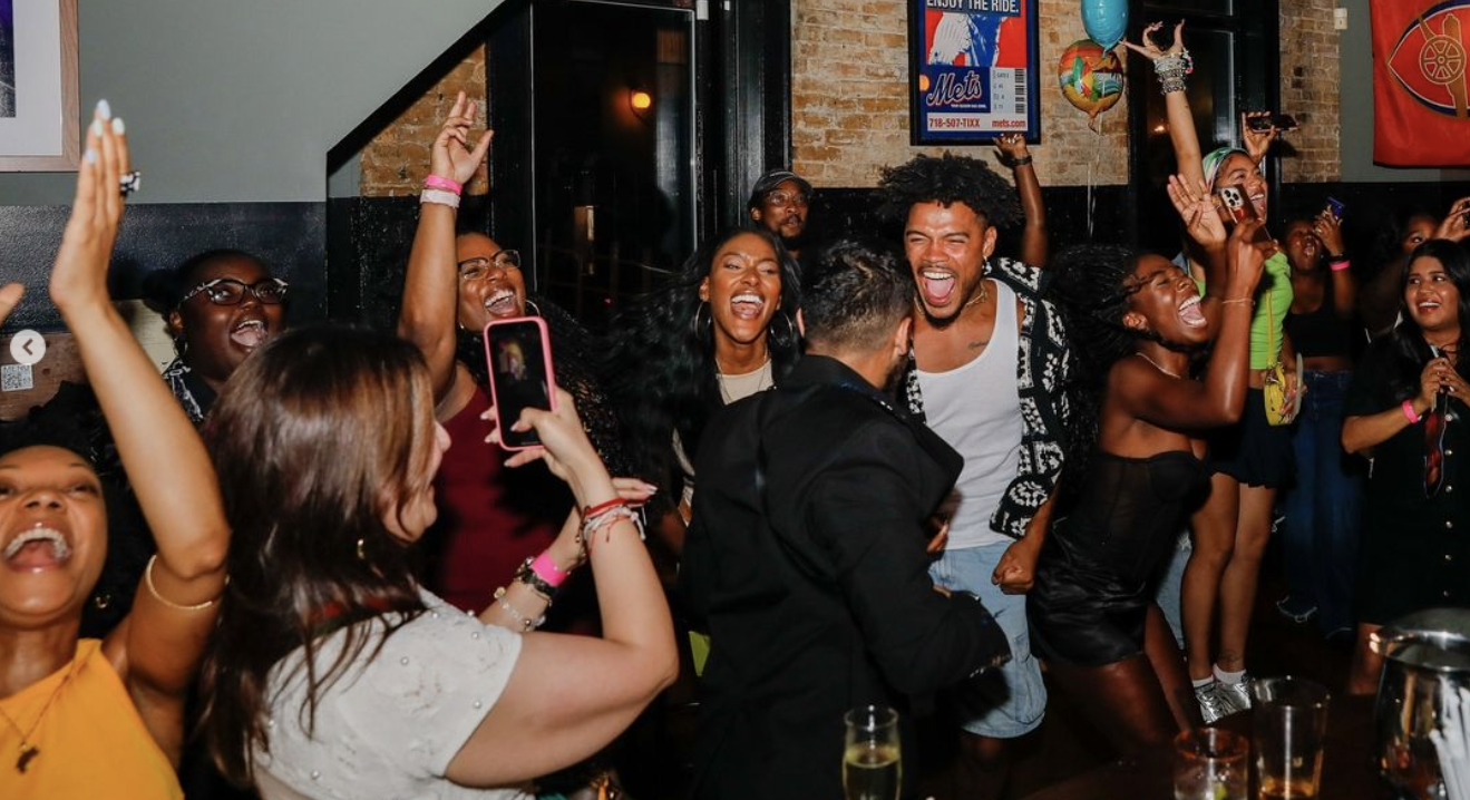 A group of people excitedly celebrate at a lively indoor party, with some holding drinks and others raising their hands in the air.