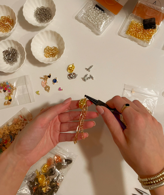 Hands assembling a gold chain using pliers, surrounded by various jewelry-making supplies like beads, chains, and hooks laid out on a white surface.