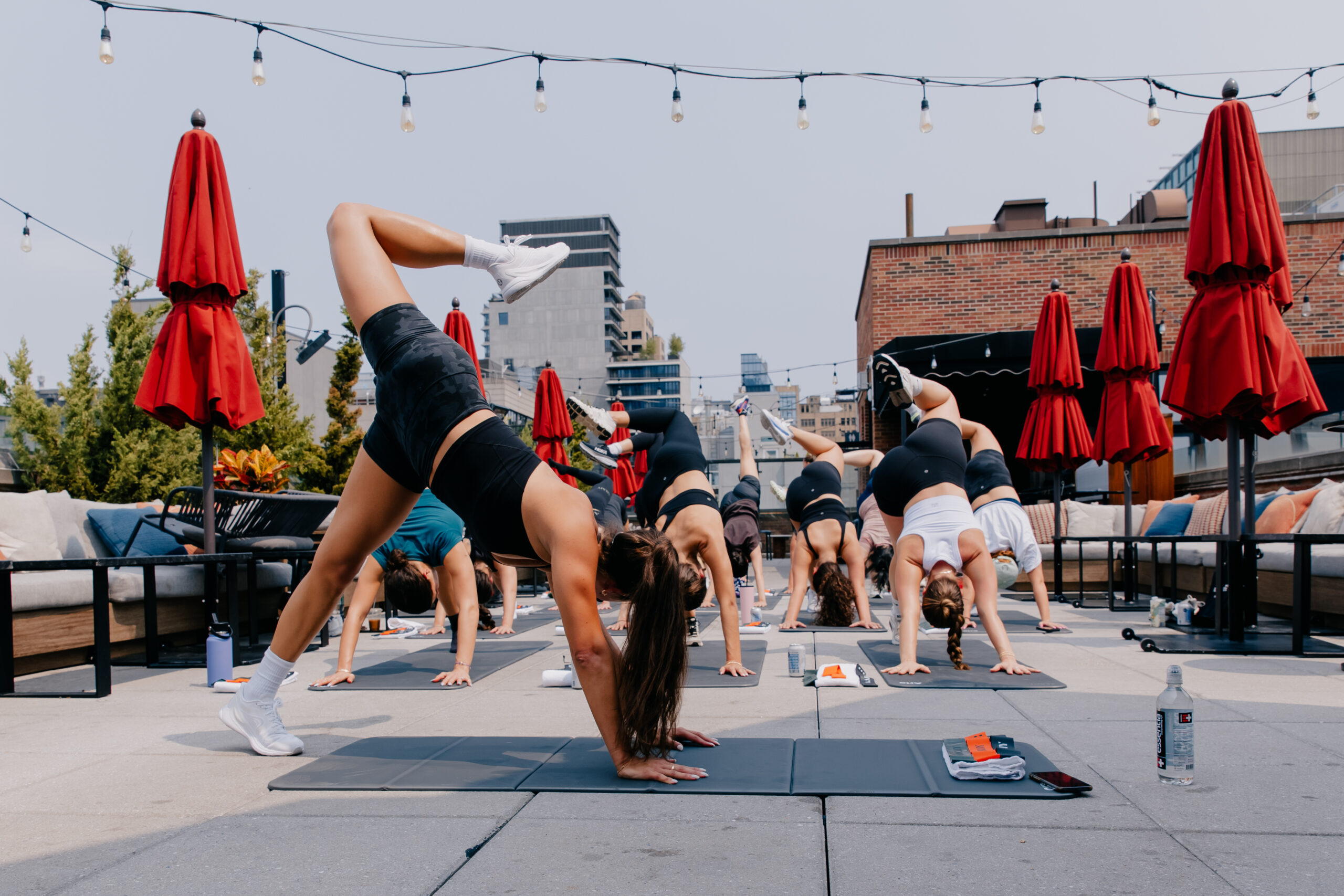 A group of people practices yoga on an outdoor rooftop, performing poses on mats under string lights and surrounded by red umbrellas.