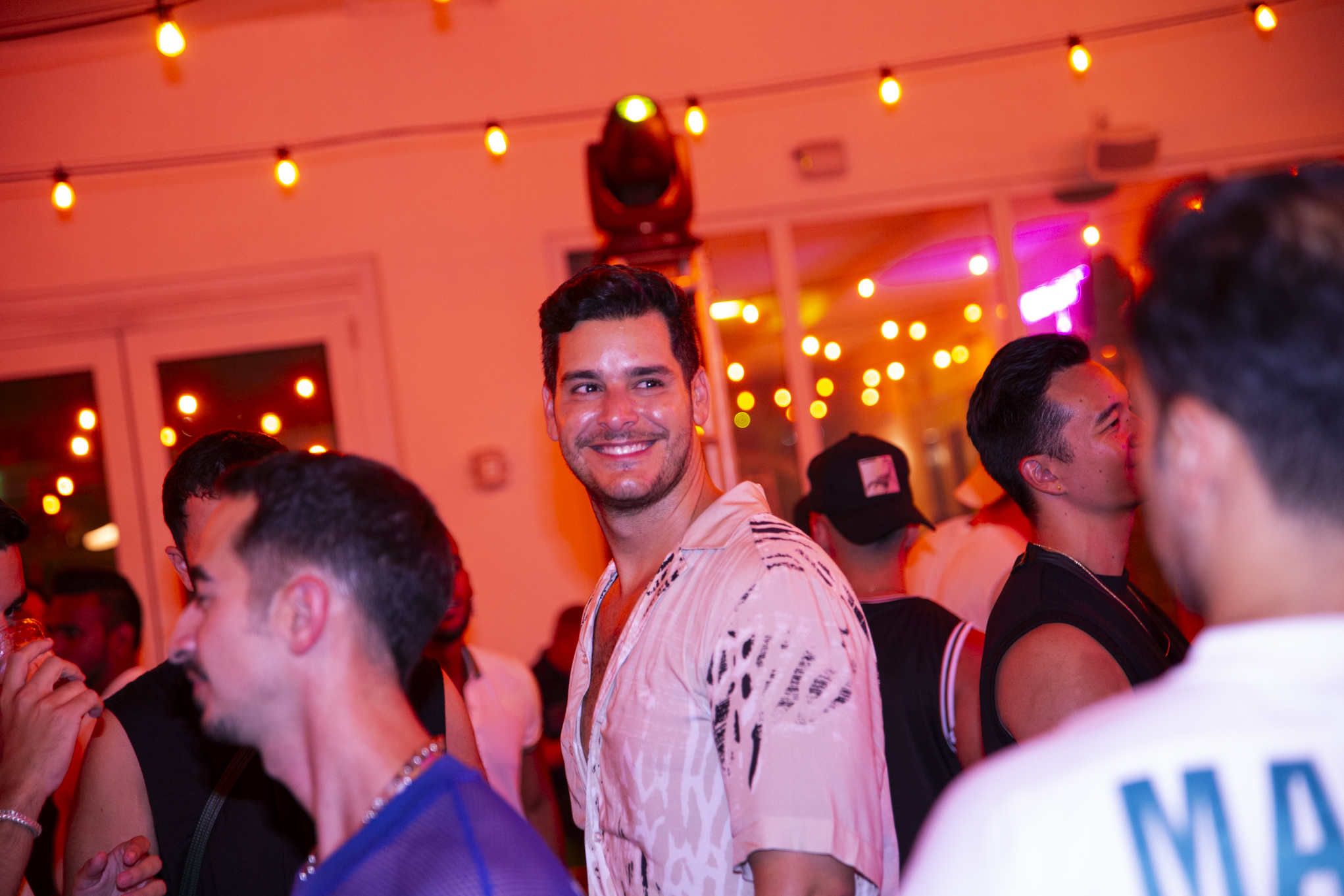 A group of people are socializing at a well-lit indoor party. One man in the center is smiling at the camera.