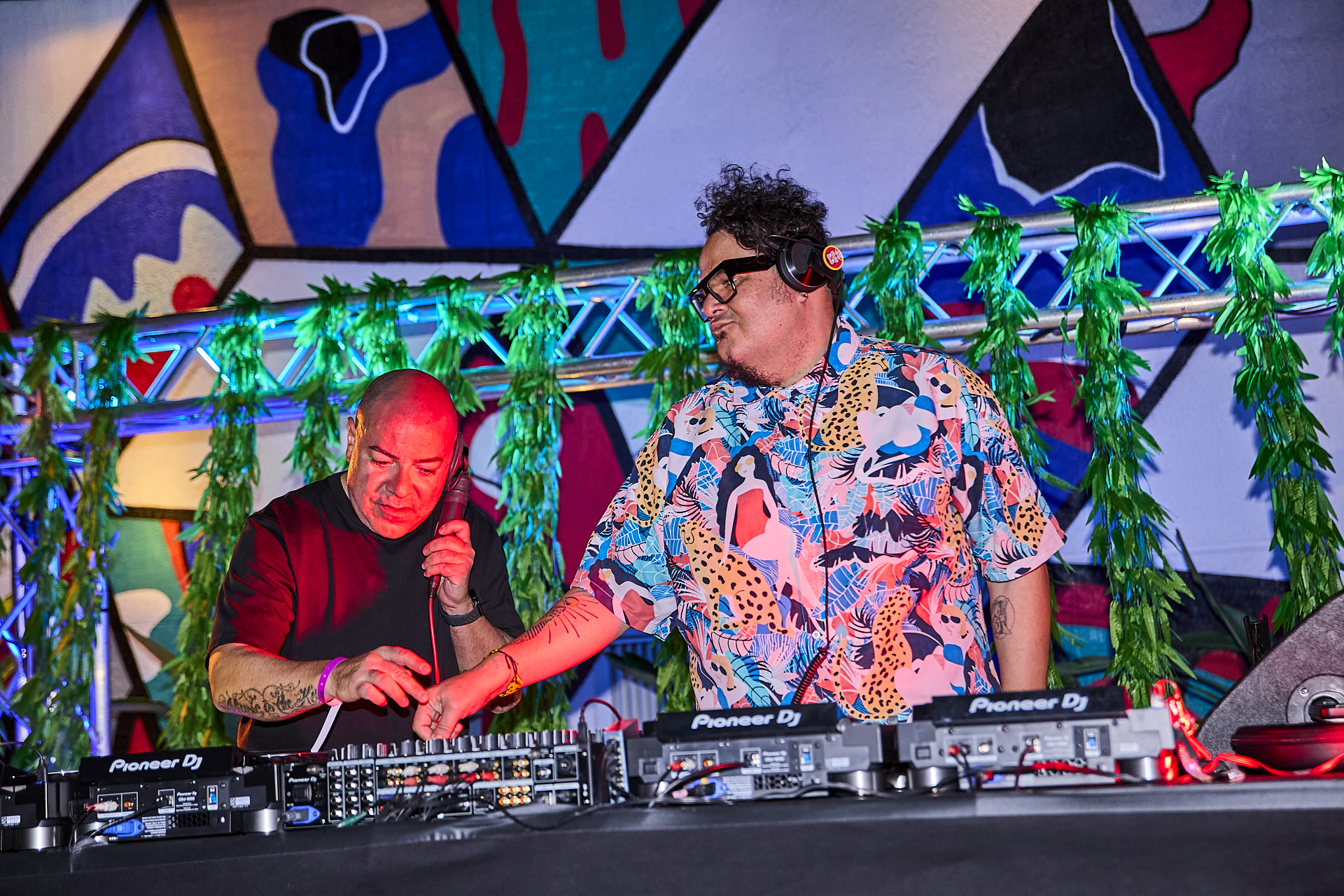 Two DJs are performing on a stage with colorful graffiti in the background. One DJ is operating the sound mixer while the other is wearing headphones and adjusting equipment.