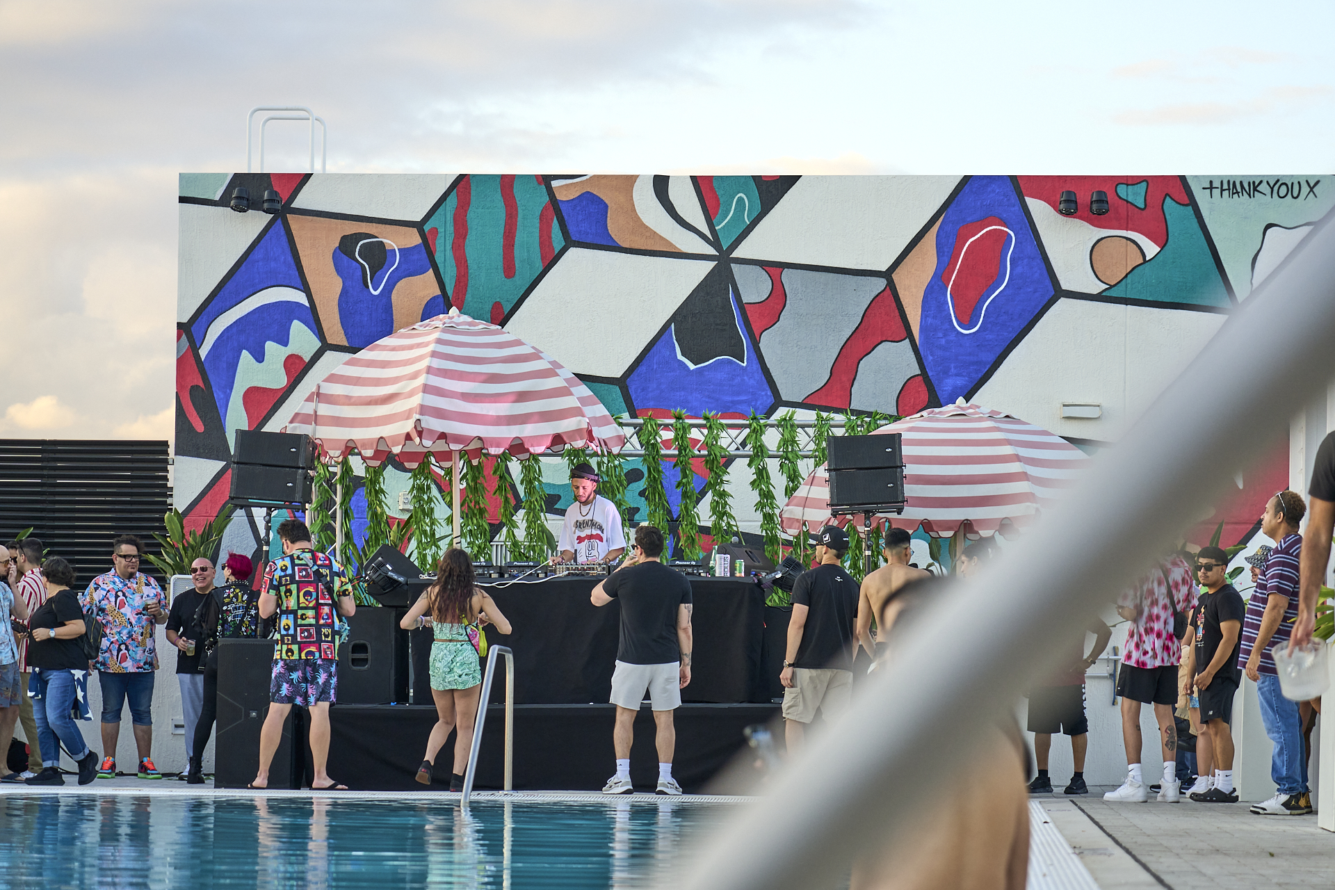 A DJ performs by a poolside with an audience gathered around. The setting features vibrant, abstract wall art and striped umbrellas.