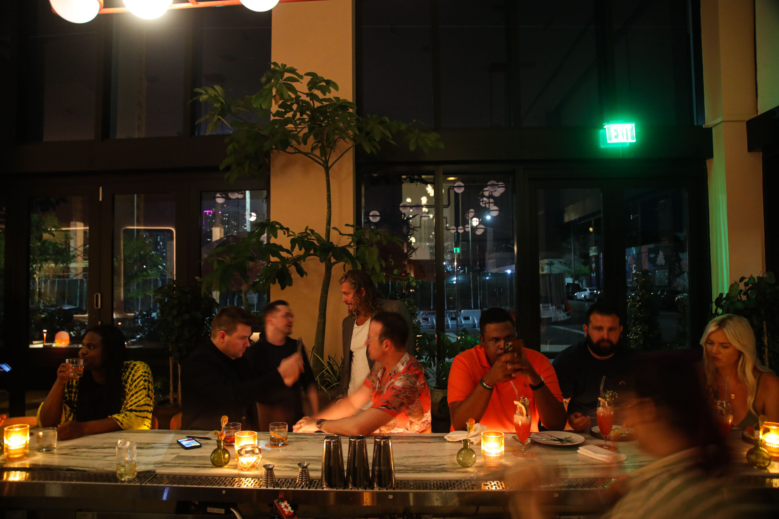 A group of people are sitting at a bar, engaged in conversation and eating. The bar is dimly lit with candles on the counter. A green exit sign is visible in the background.