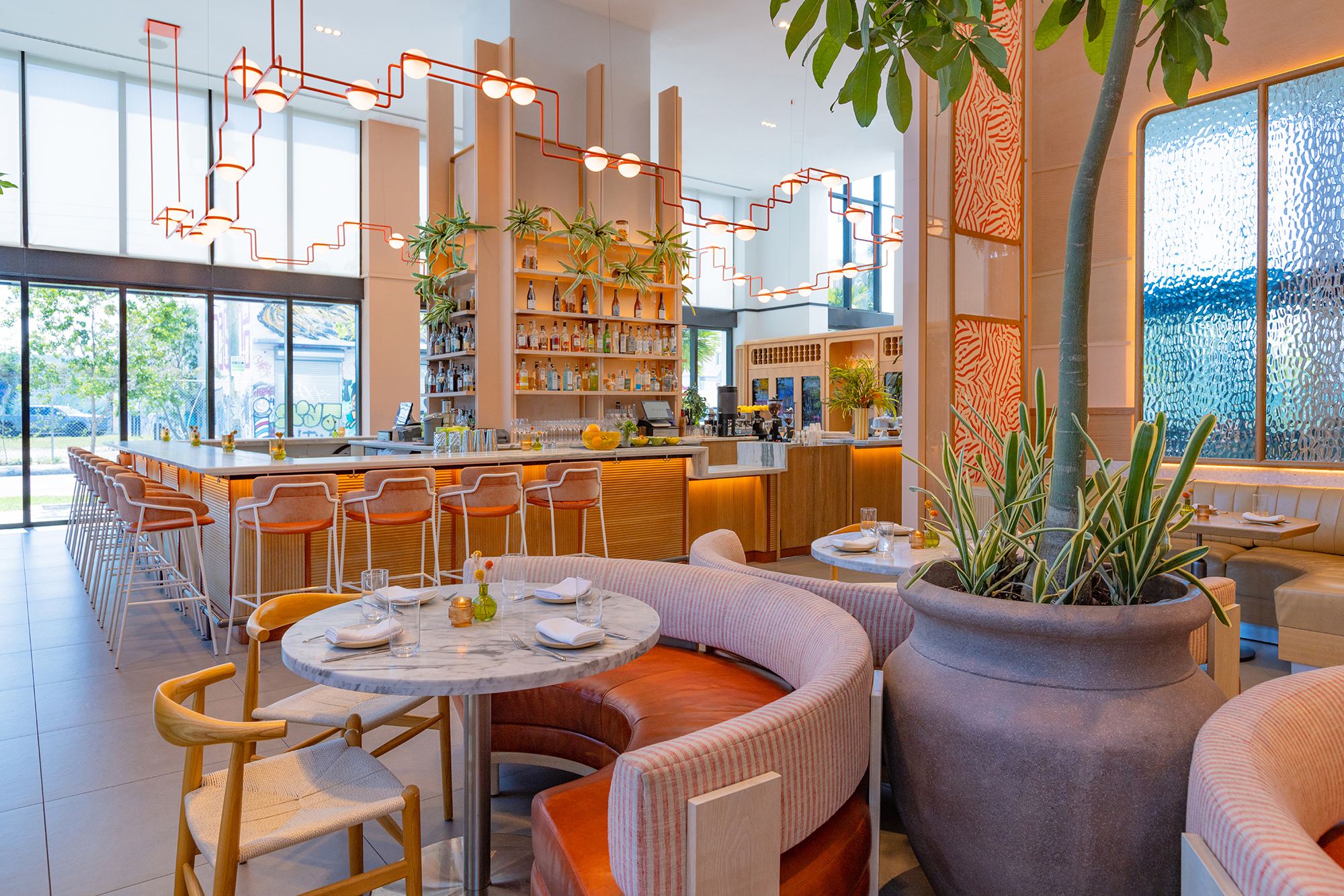 Spacious modern restaurant with a bar counter, high chairs, large potted plant, round tables, cushioned seating, and decorative lighting. Large windows provide natural light.