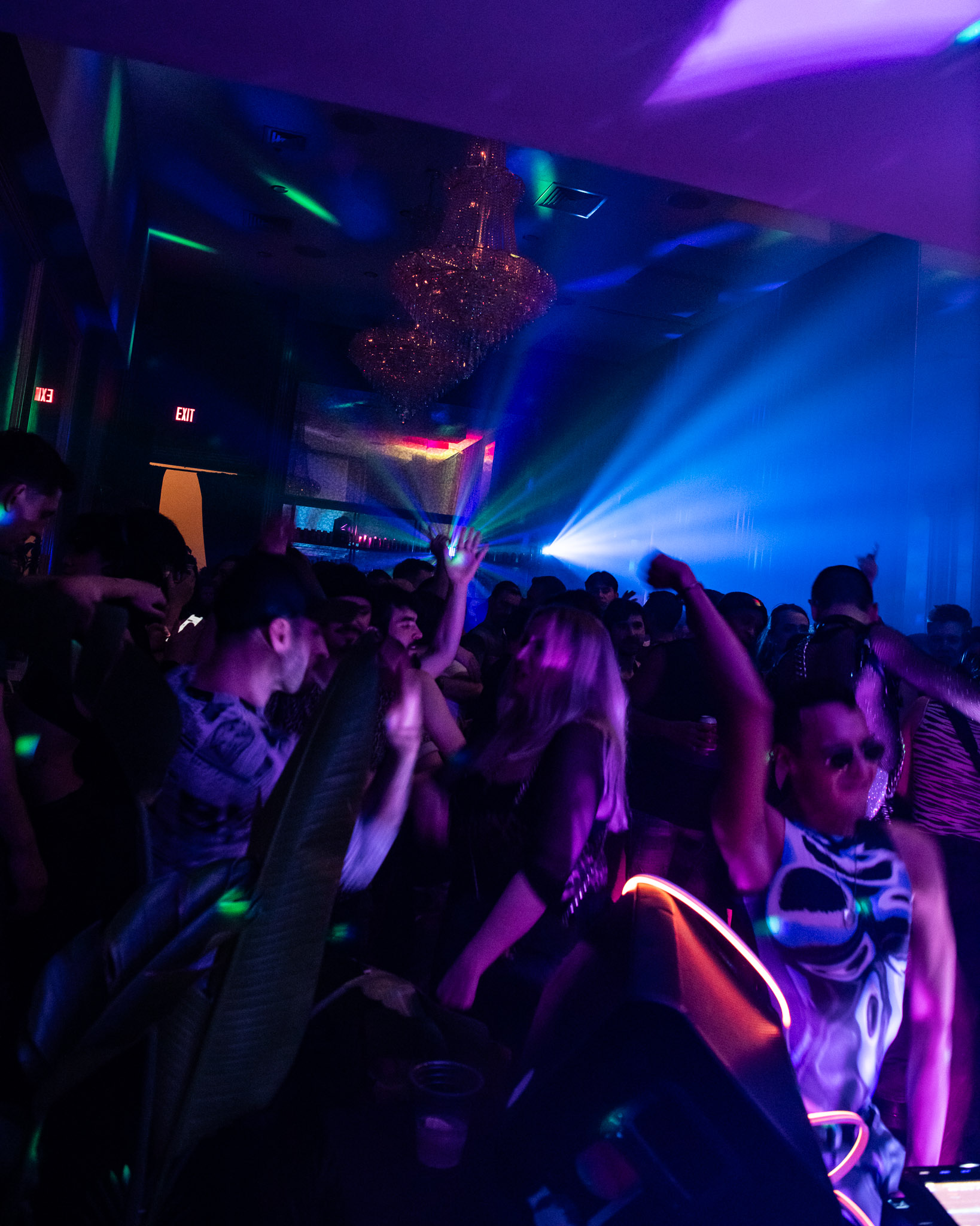 A crowded nightclub dance floor with colorful lights, people dancing energetically, and a chandelier overhead.