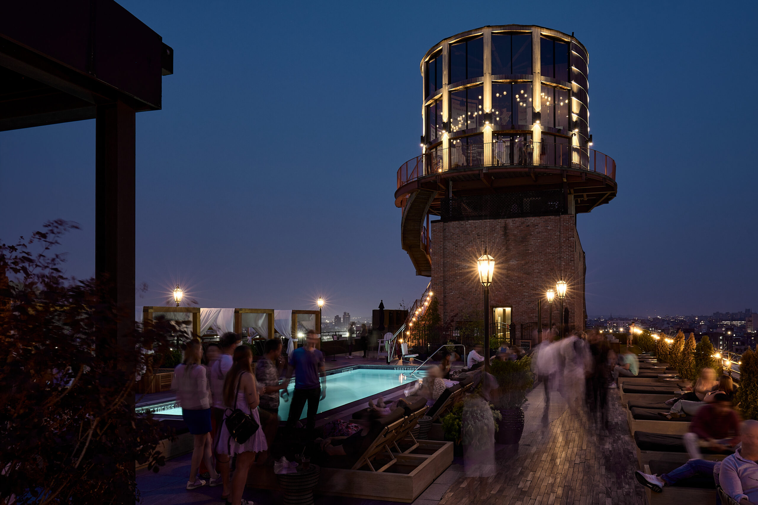 A rooftop bar at dusk, featuring a modern tower structure, a swimming pool with people around, and city lights in the background.