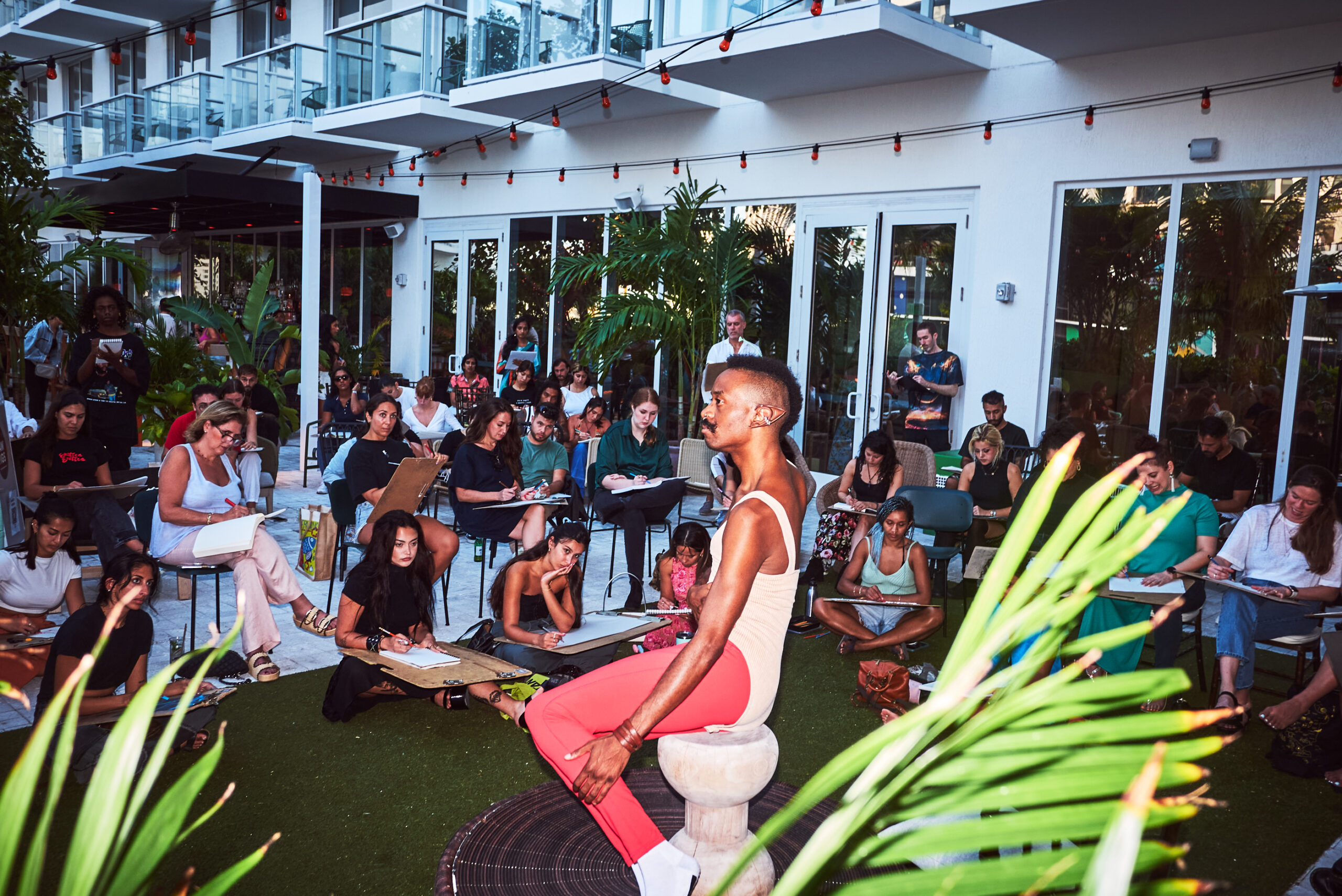 A group of people sitting outside, drawing a person posing on a pedestal. The setting is a patio with greenery and a building in the background.