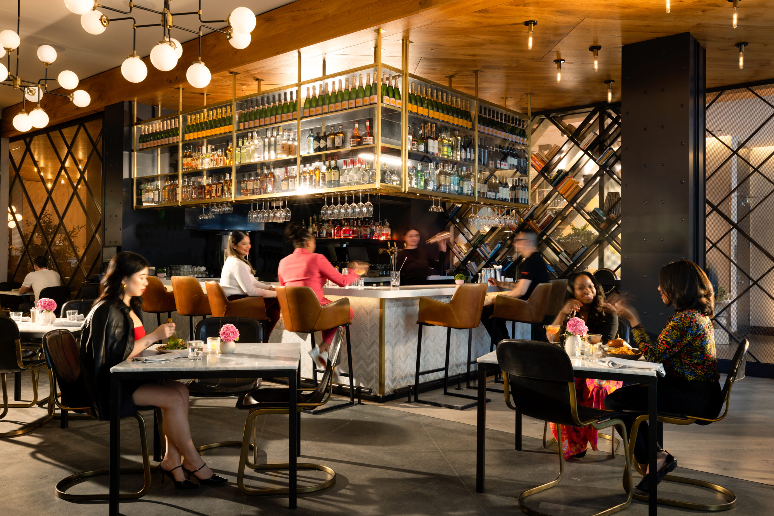 People sitting and standing at a modern bar with shelves of liquor bottles and pendant lights. Some are at tables, and others are at the bar where bartenders are serving drinks.